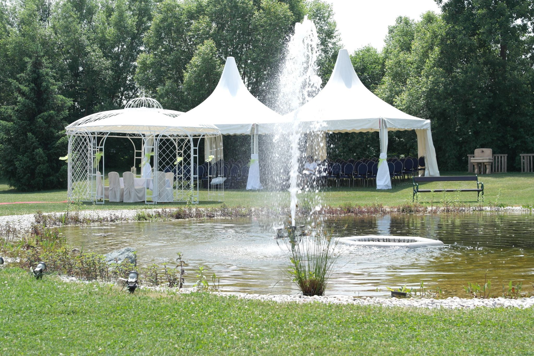 A wedding pavilion and tents next to a pond with a fountain in a garden.