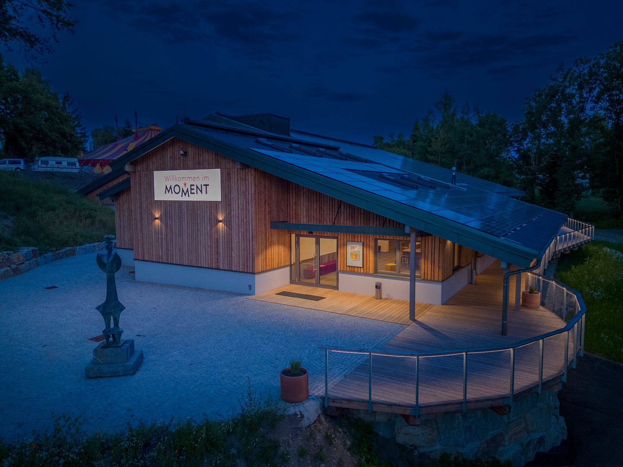 A modern building with wooden cladding and solar panels on the roof, illuminated at dusk. A sign with the inscription 'Welcome to MOMENT' is visible.