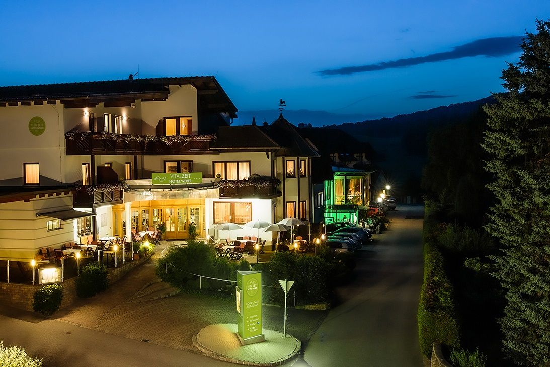 Exterior view of the Vitalzeit Hotel Weber at night, illuminated with warm light, surrounded by trees and parked cars.