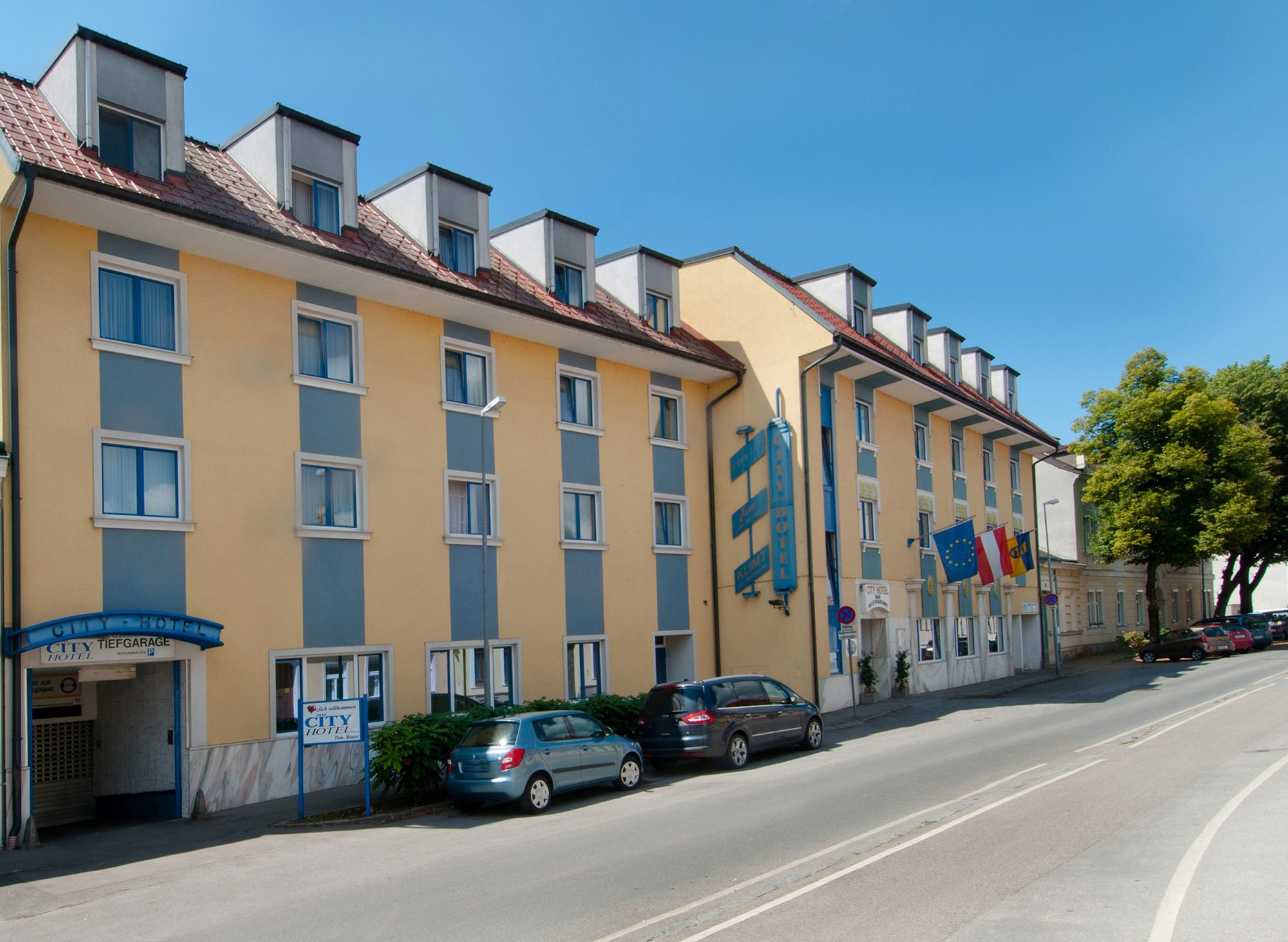 Exterior view of a yellow-blue hotel with several windows and parked cars in front of it.