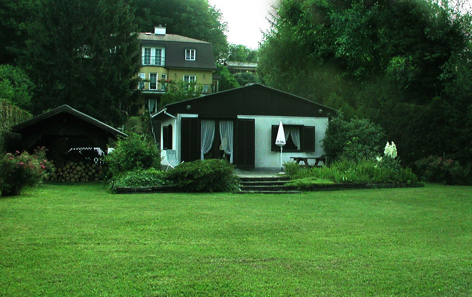 A small house with a garden and terrace, surrounded by trees and a larger building in the background.