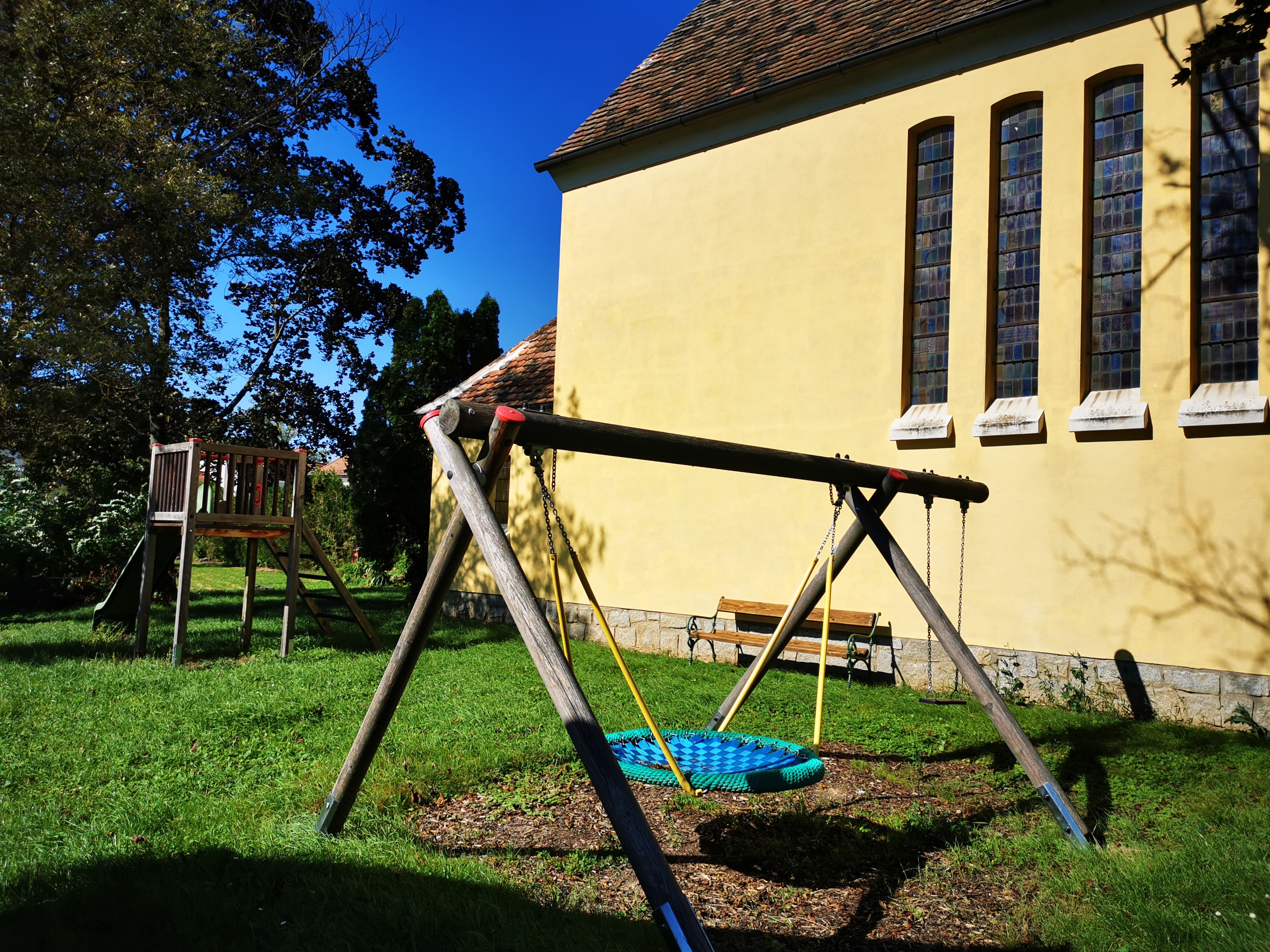 Playground with swing and slide next to a yellow chapel with high windows.