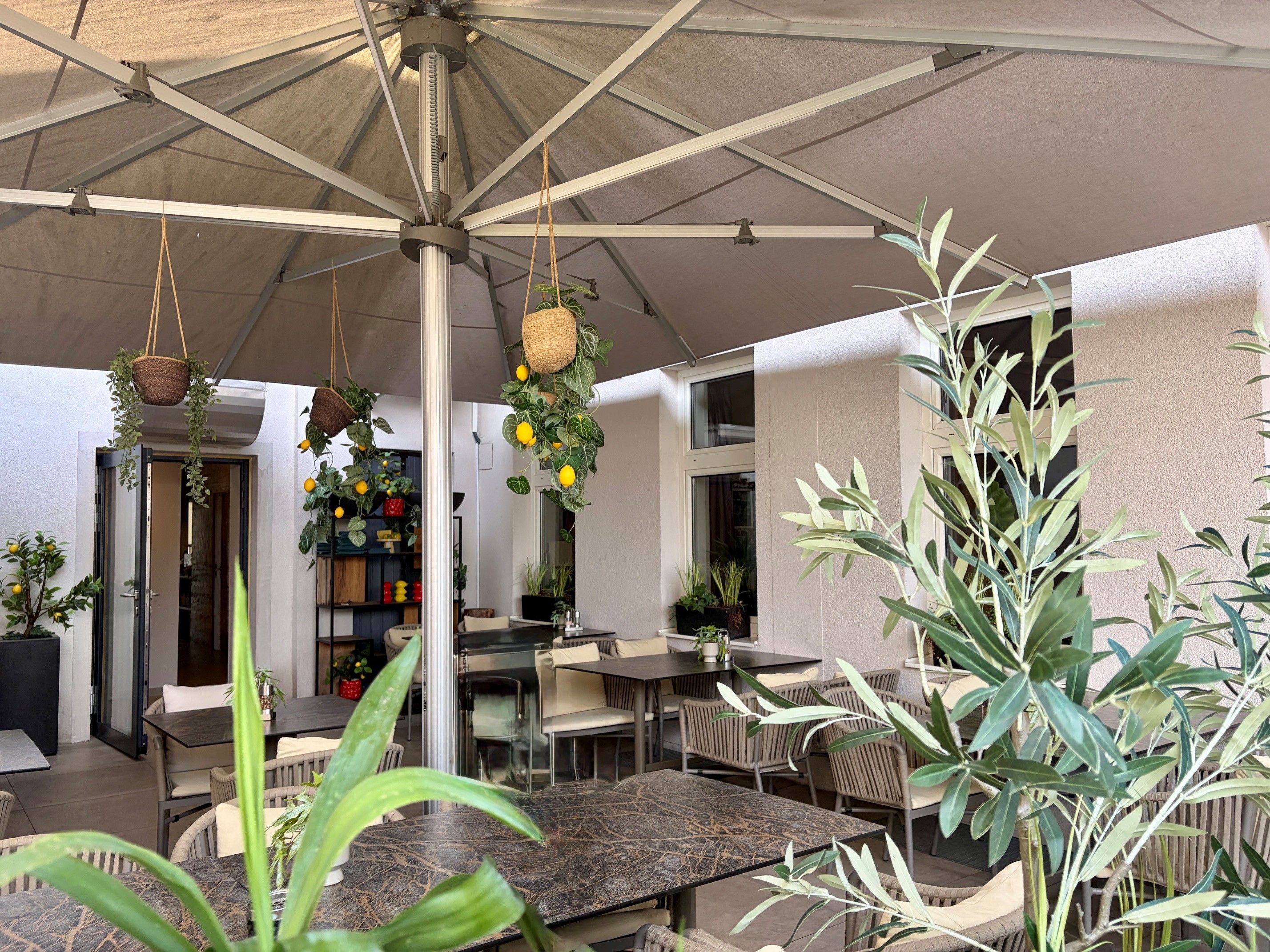 A cozy outdoor dining area with a parasol, hanging plants, and tables, surrounded by greenery and decorations.