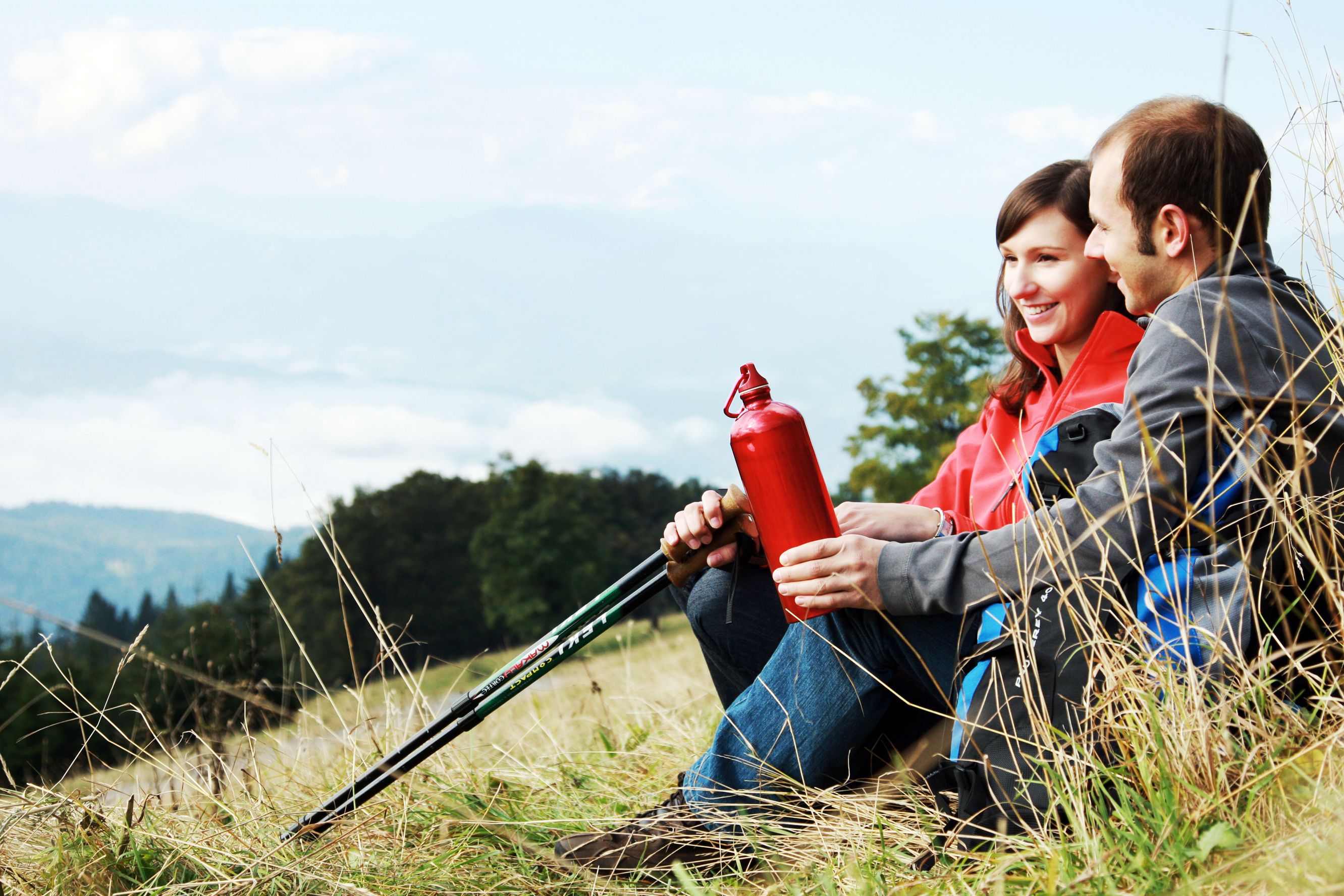 A couple sits on a meadow with hiking poles and a red water bottle, mountains and clouds in the background.