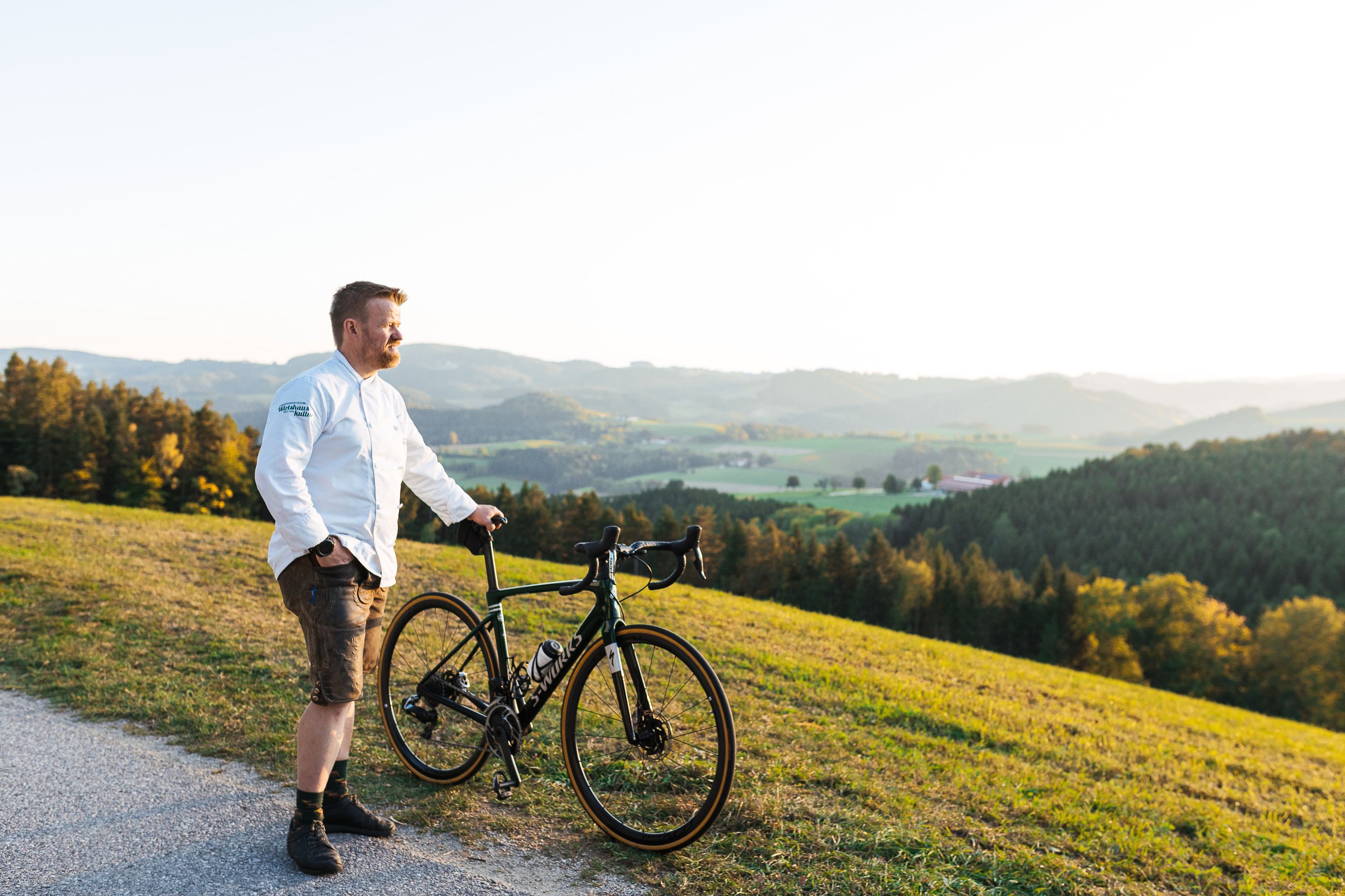 A man in a white shirt stands with a bicycle on a hill overlooking a green landscape.