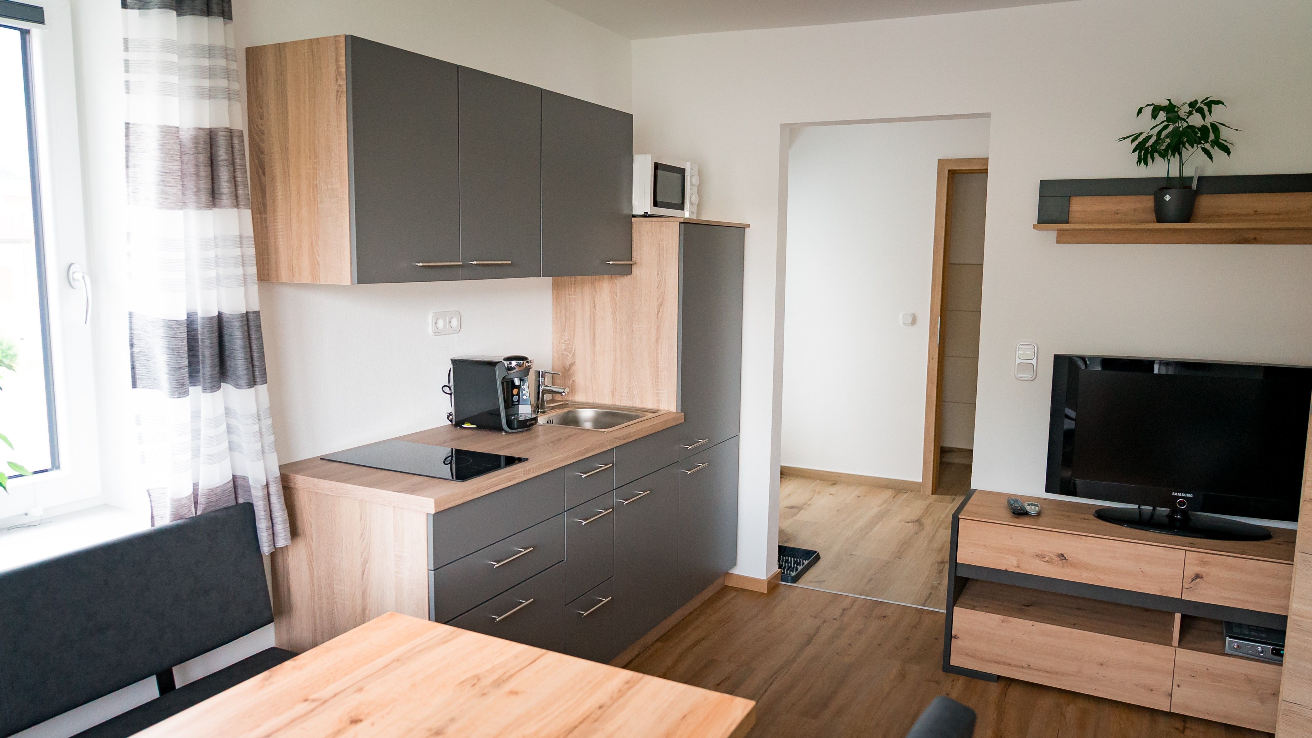Modern kitchen with gray cabinets, wooden worktop, coffee machine, sink, microwave and TV on wooden shelf.