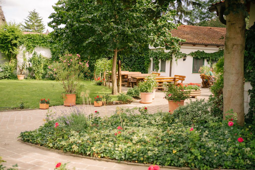 A well-tended garden with lawn, flower beds and terracotta pots. In the background, a house with a terrace and wooden tables.