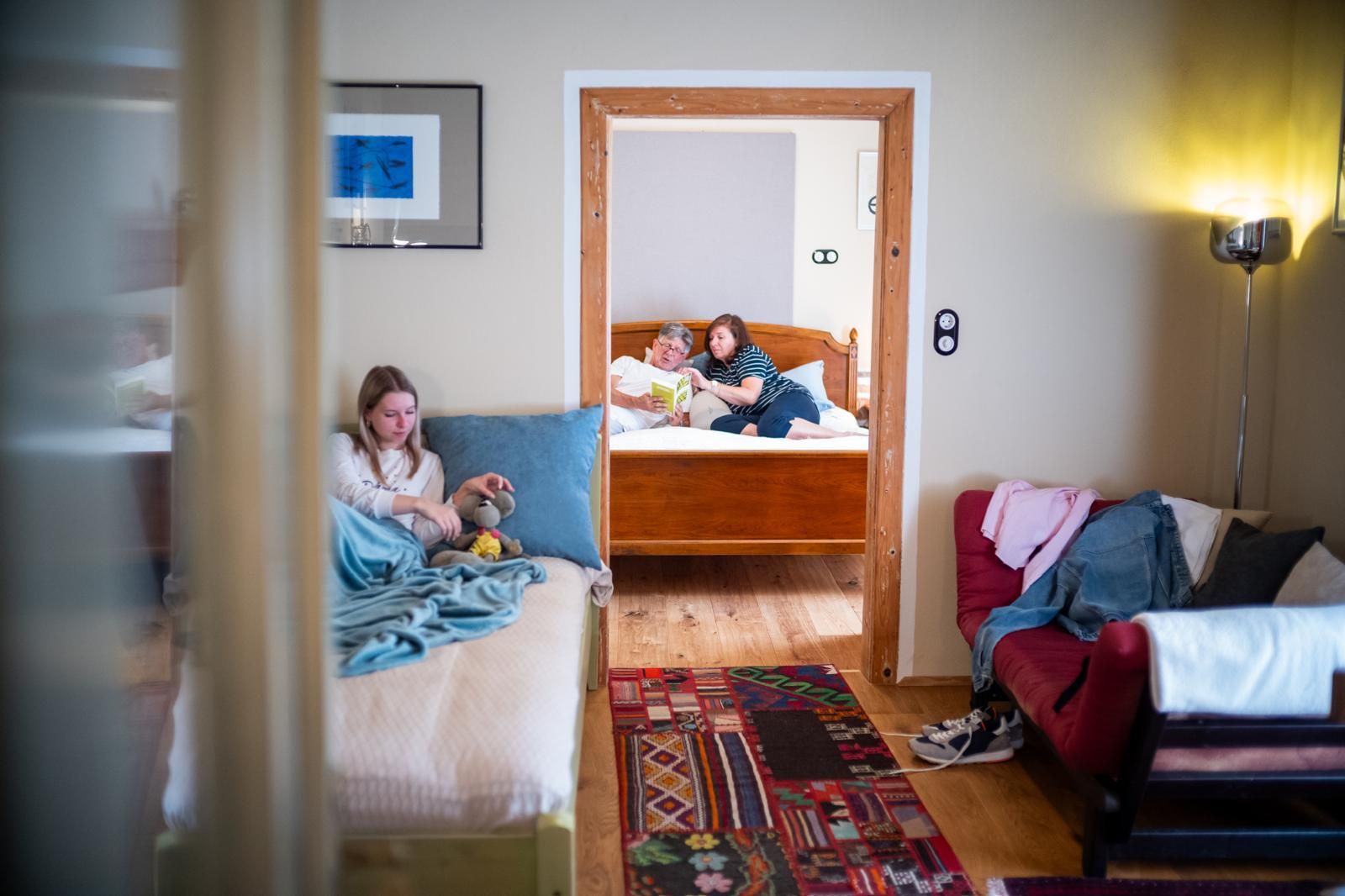 Interior view of a vacation apartment with two rooms. In the foreground a girl is sitting on a bed, in the background two people are reading on another bed.