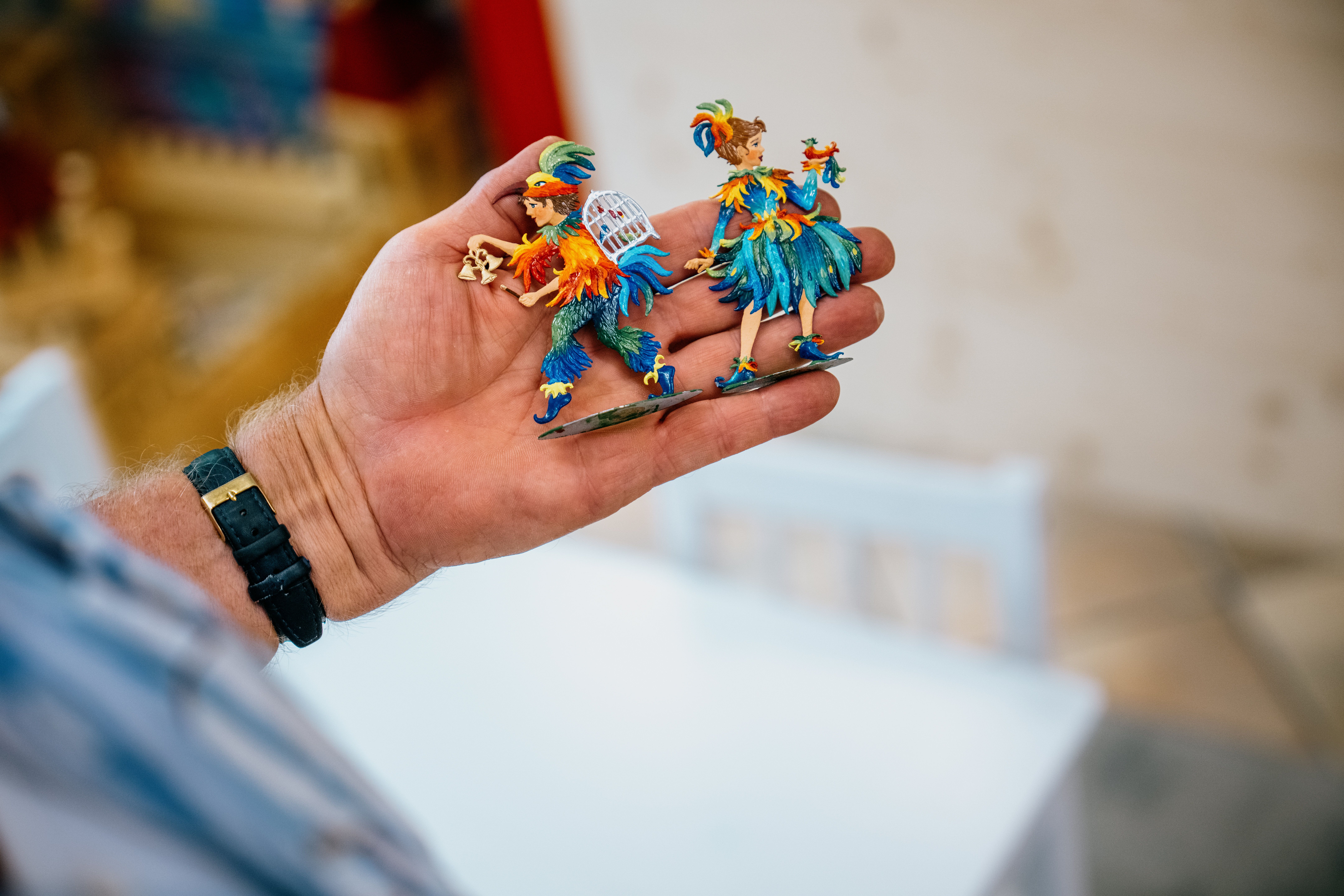 A hand is holding two colorful pewter figurines dressed in parrot costumes.