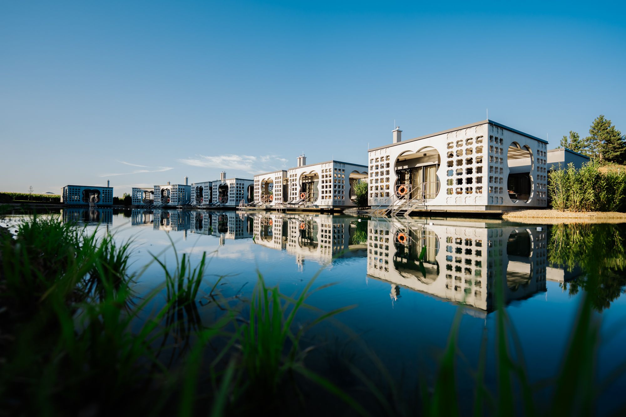 Modern villas on the water with reflections, surrounded by nature.