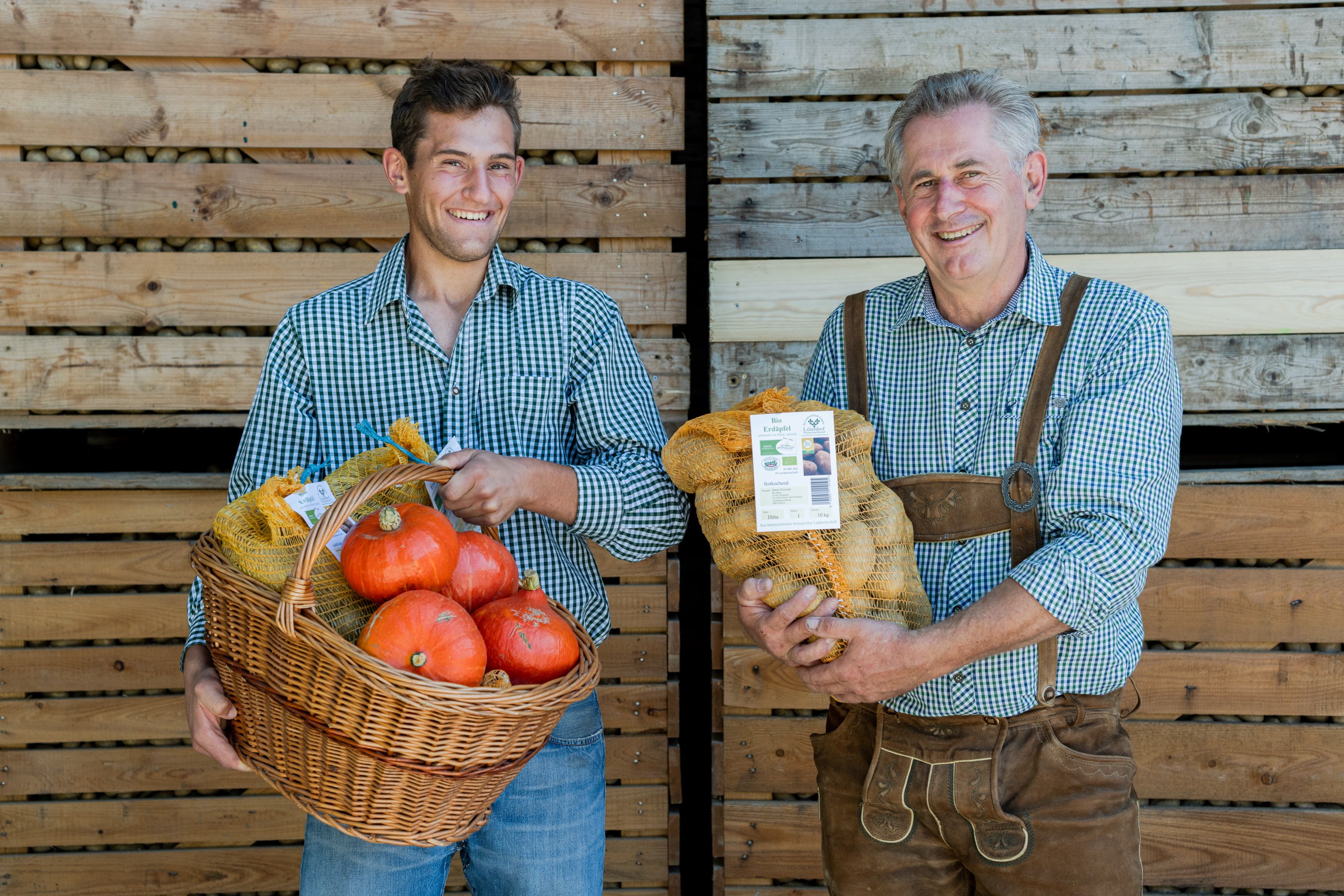 Two smiling men in checked shirts hold a basket of pumpkins and a sack of potatoes in front of a wooden wall.