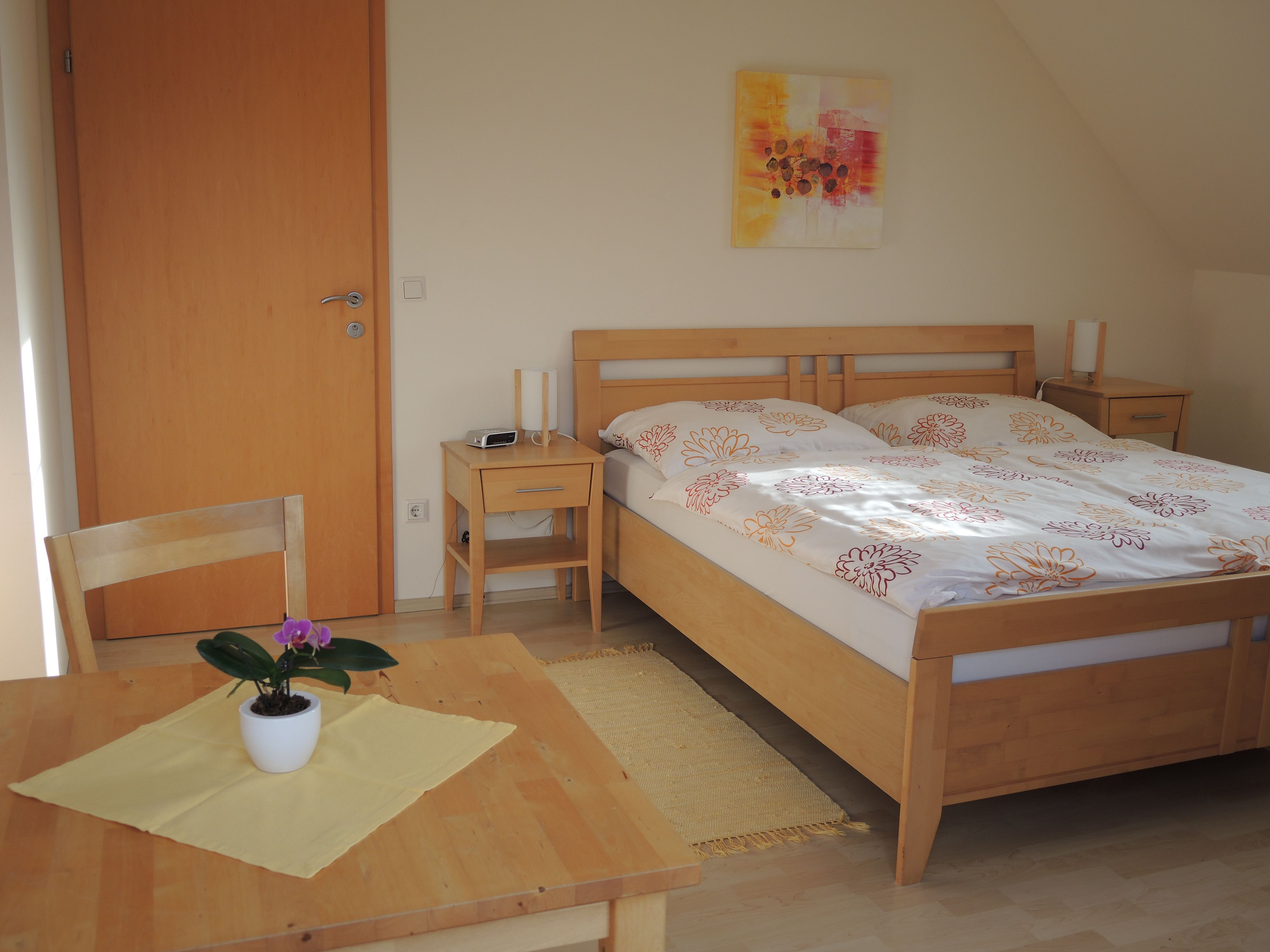 A guest room with a double bed, two bedside tables, a table and chair and a potted plant.
