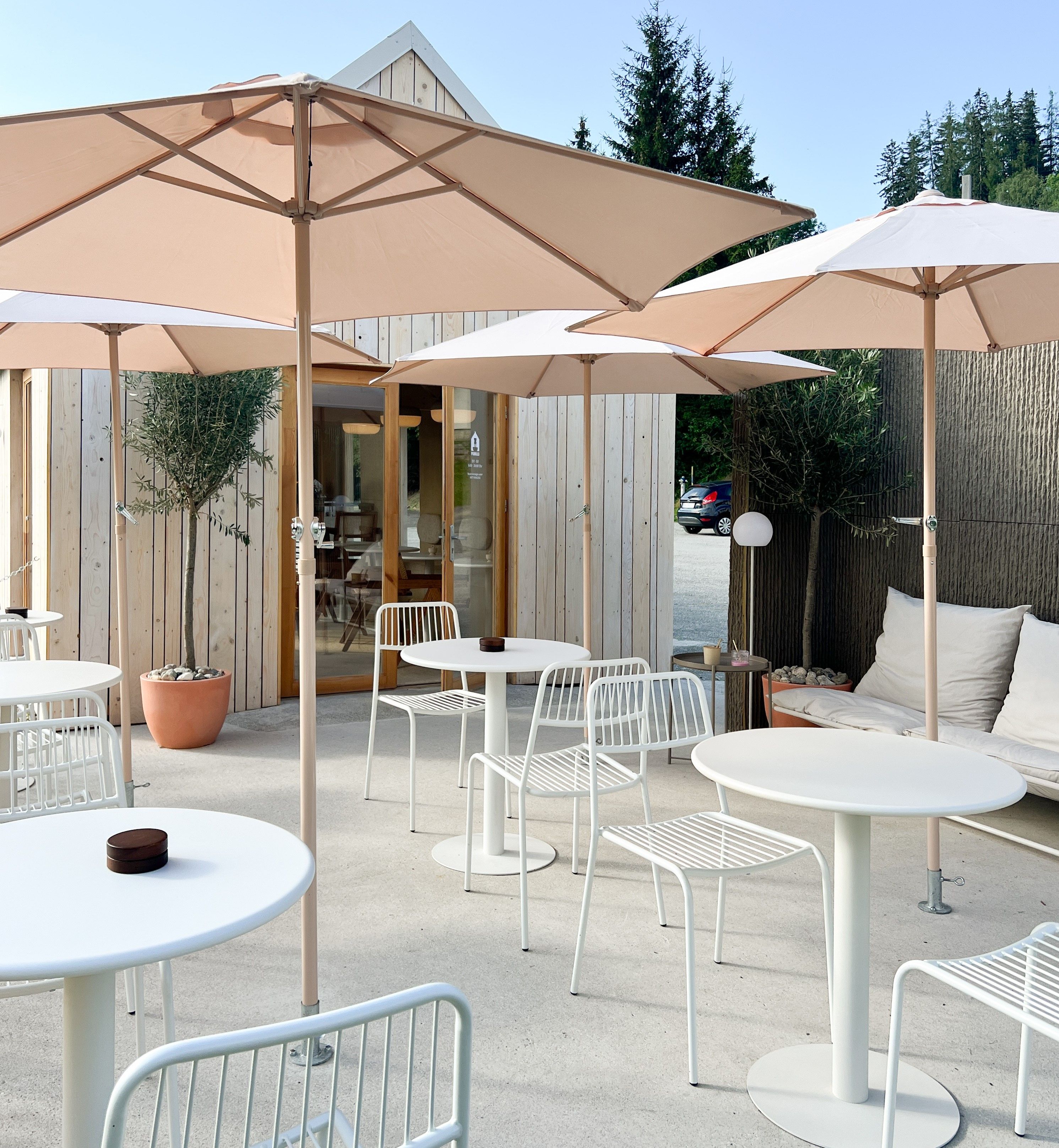 Modern terrace with white tables and chairs, parasols and wooden hut in the background.