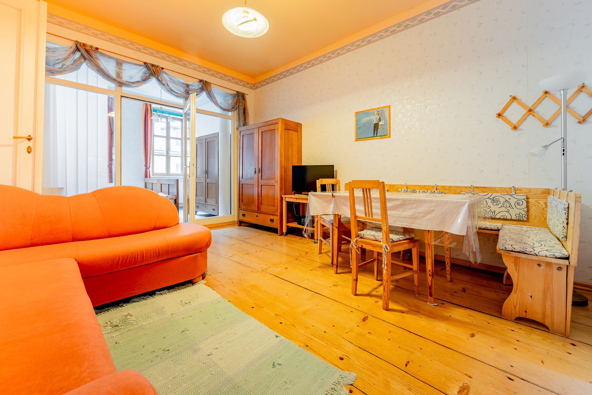 A living room with an orange sofa, wooden table and chairs, wooden floor and a cupboard. A picture hangs on the wall.
