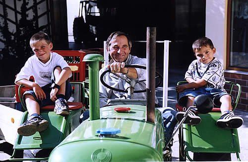 A man sits on a green tractor, flanked by two boys on toy tractors.