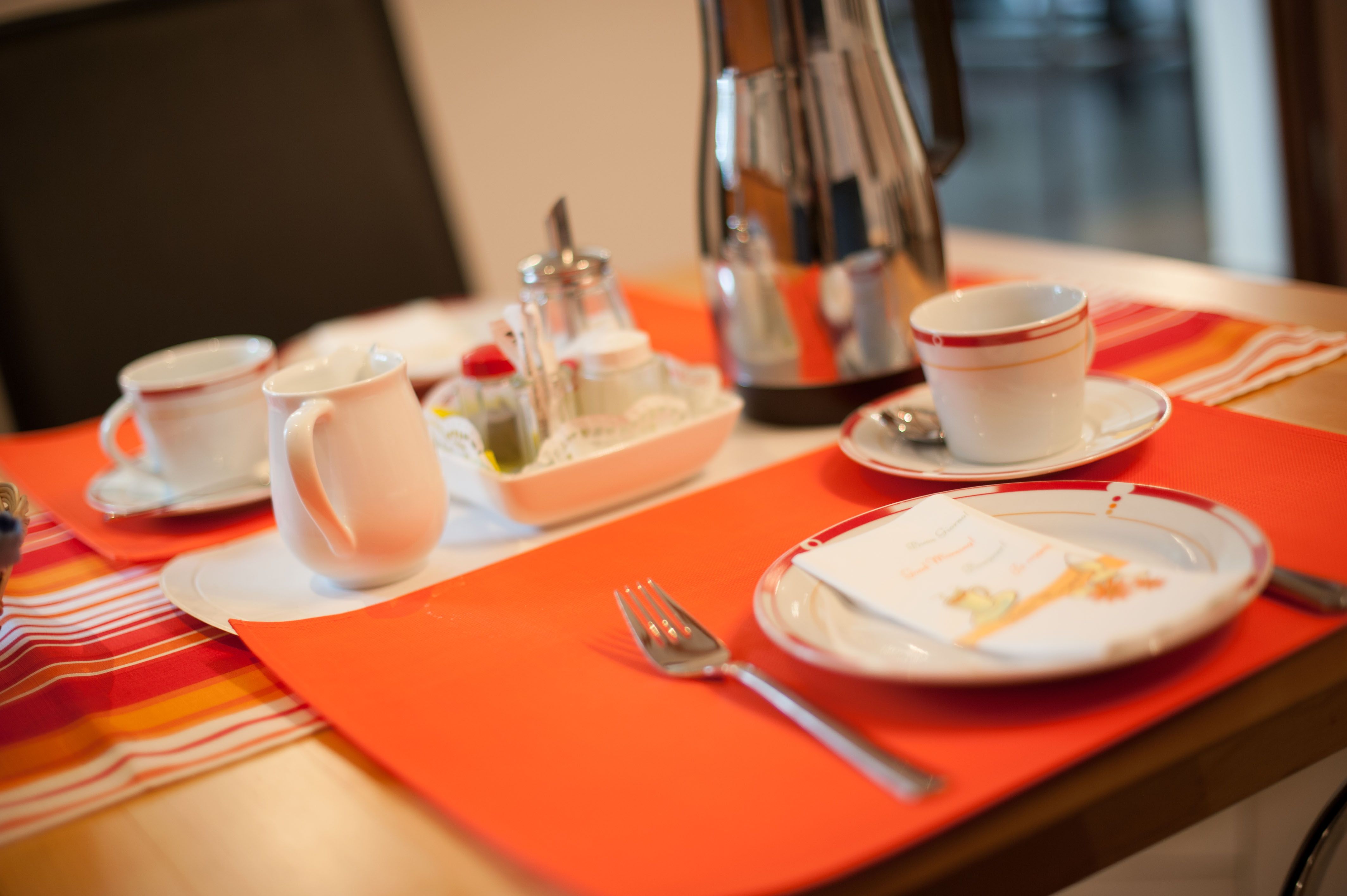 Breakfast table with cups, plates, cutlery and coffee pot on an orange place mat.