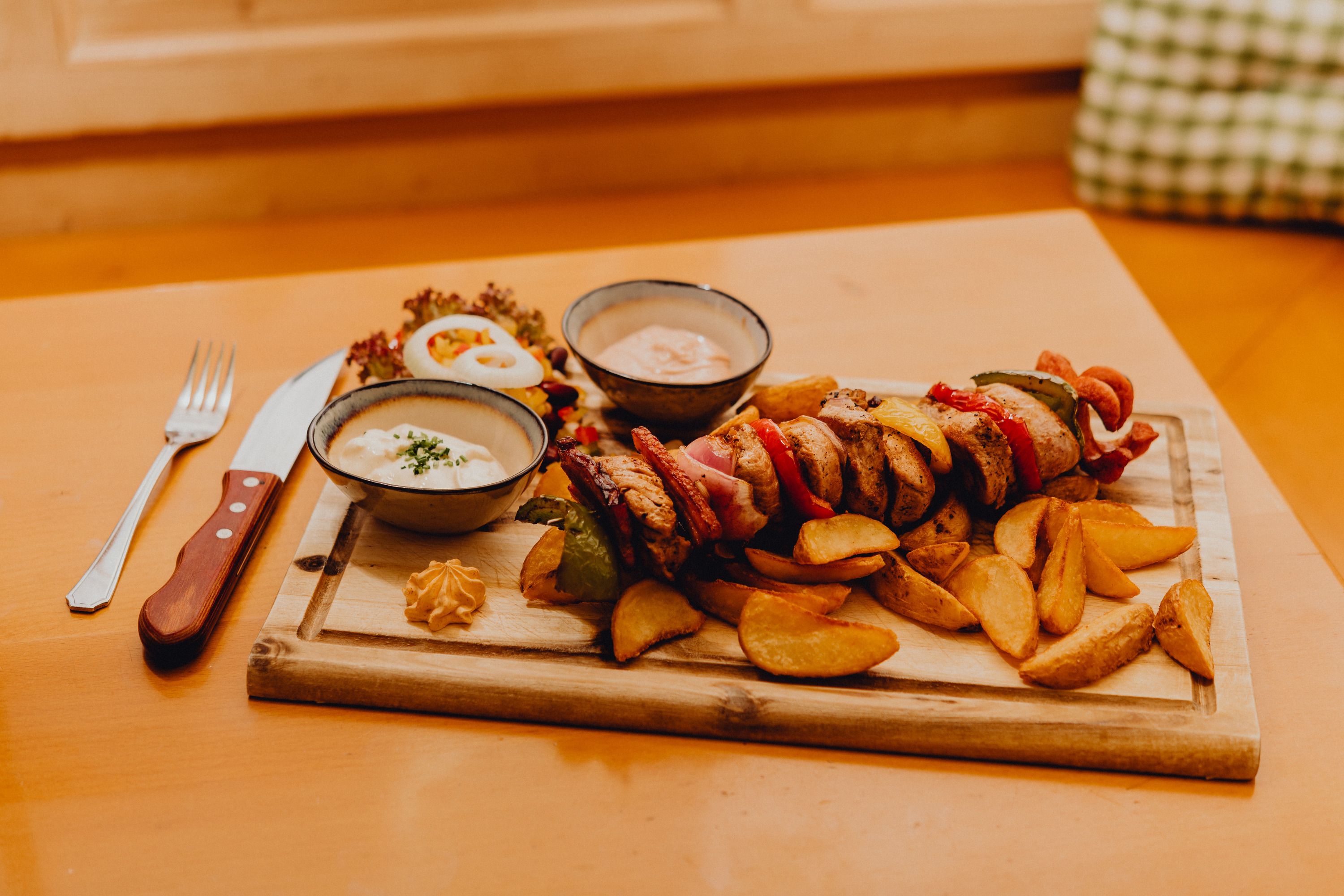 A wooden board with a meat skewer, potato wedges, two sauce bowls and salad on a table.