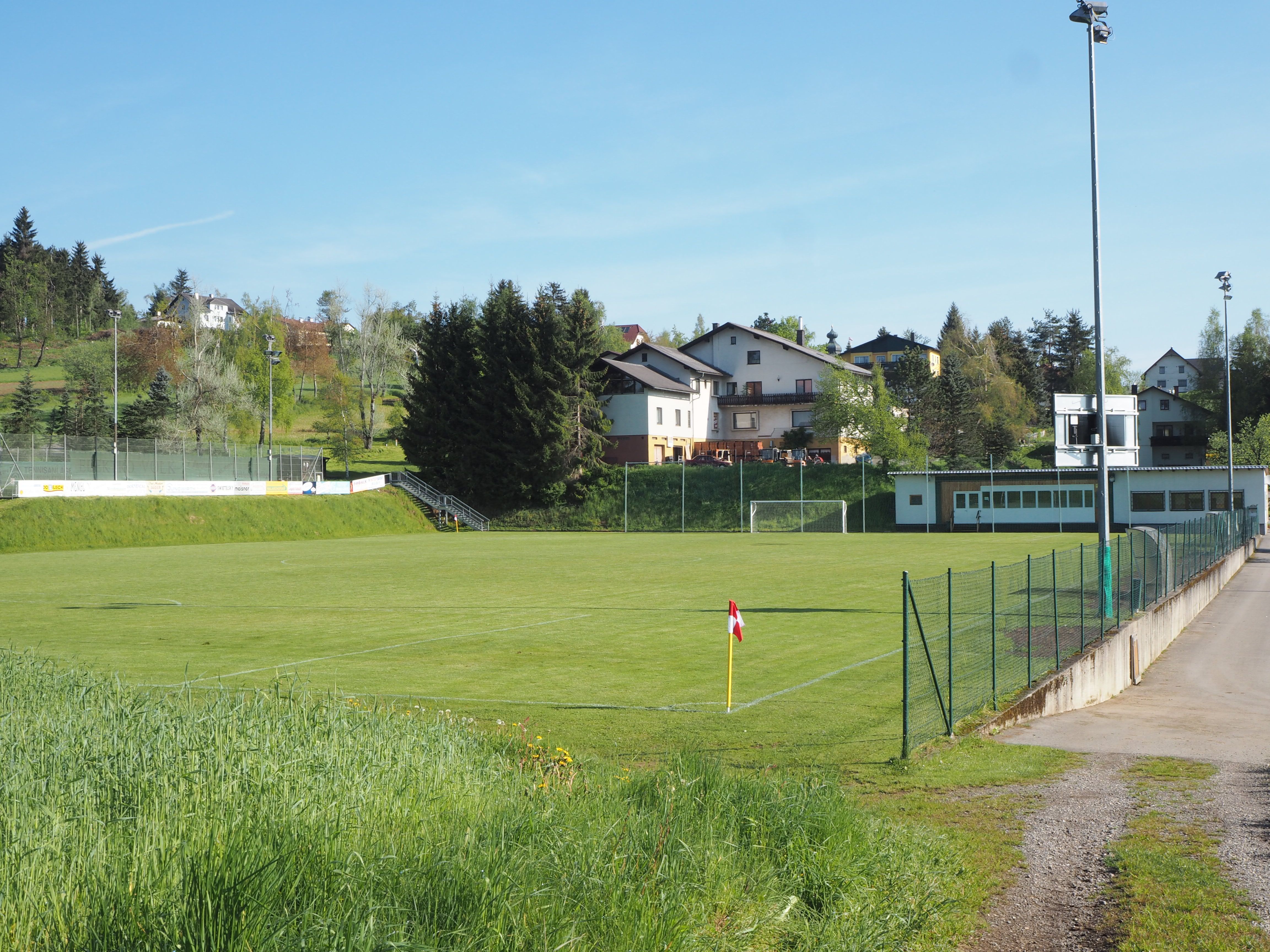 An empty soccer pitch with surrounding buildings and trees on a sunny day.