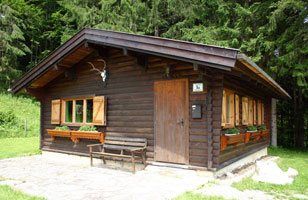 A small, rustic wooden hunting lodge with shutters and antlers on the façade, surrounded by forest.