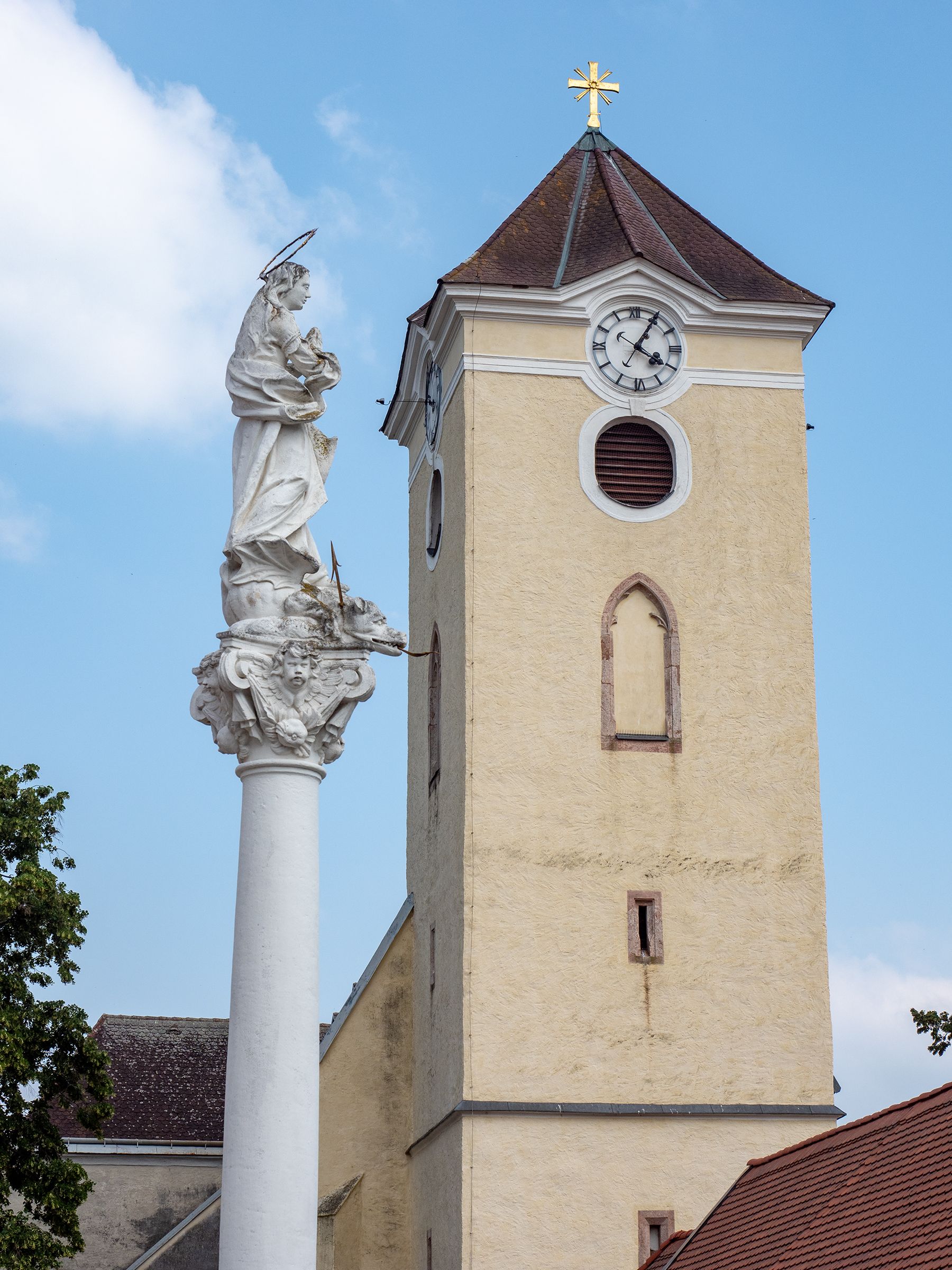 Church tower with clock and Marian column in Schrattenthal against a blue sky.