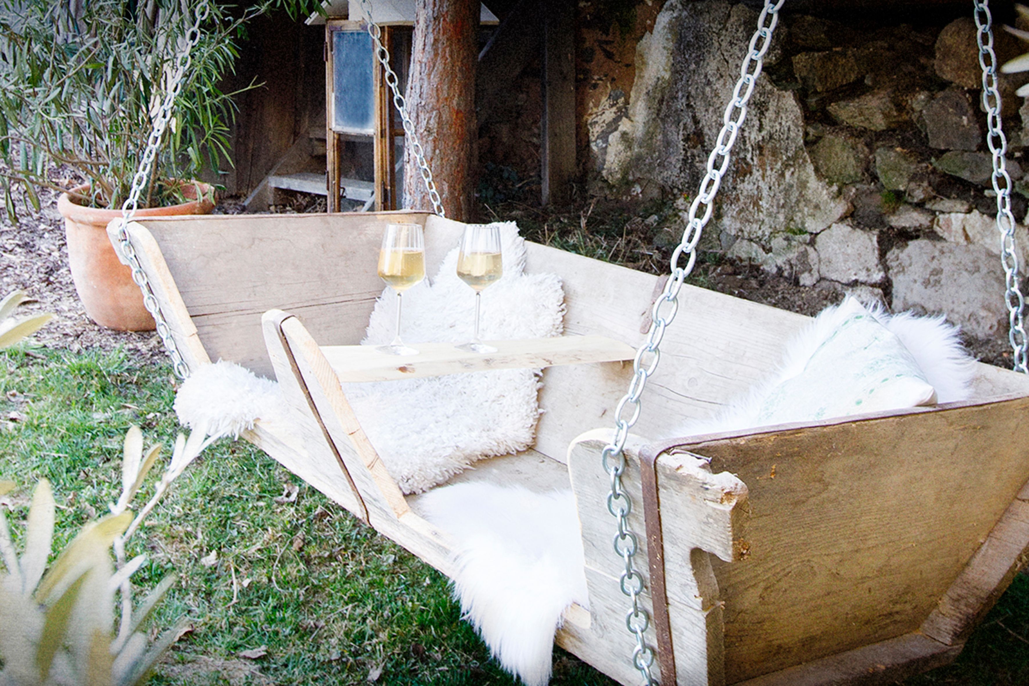 A wooden swing in the garden with a pillow and two glasses of white wine on a table.