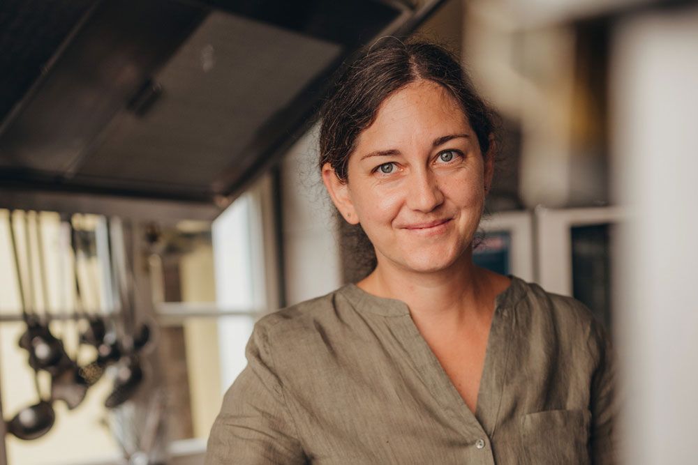 A woman in a kitchen, smiling, with brown hair and a green shirt.
