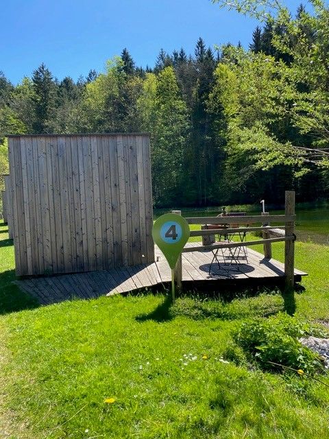A small wooden cabin with a patio and a table, nestled in the countryside and surrounded by trees, located right next to the pond. 