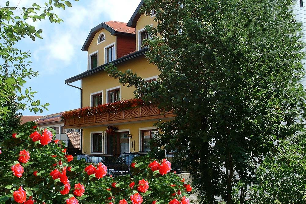 Yellow hotel building with balcony, surrounded by blooming red roses and trees.