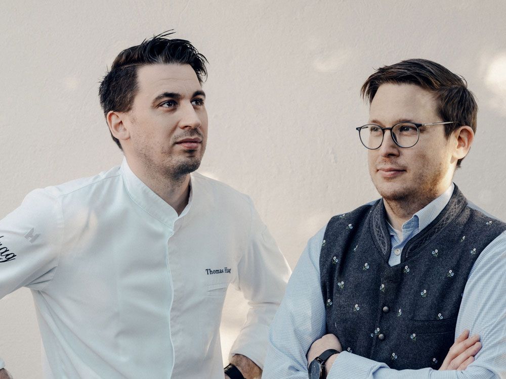 Two men are standing next to each other, one wearing a chef's jacket, the other a traditional vest.