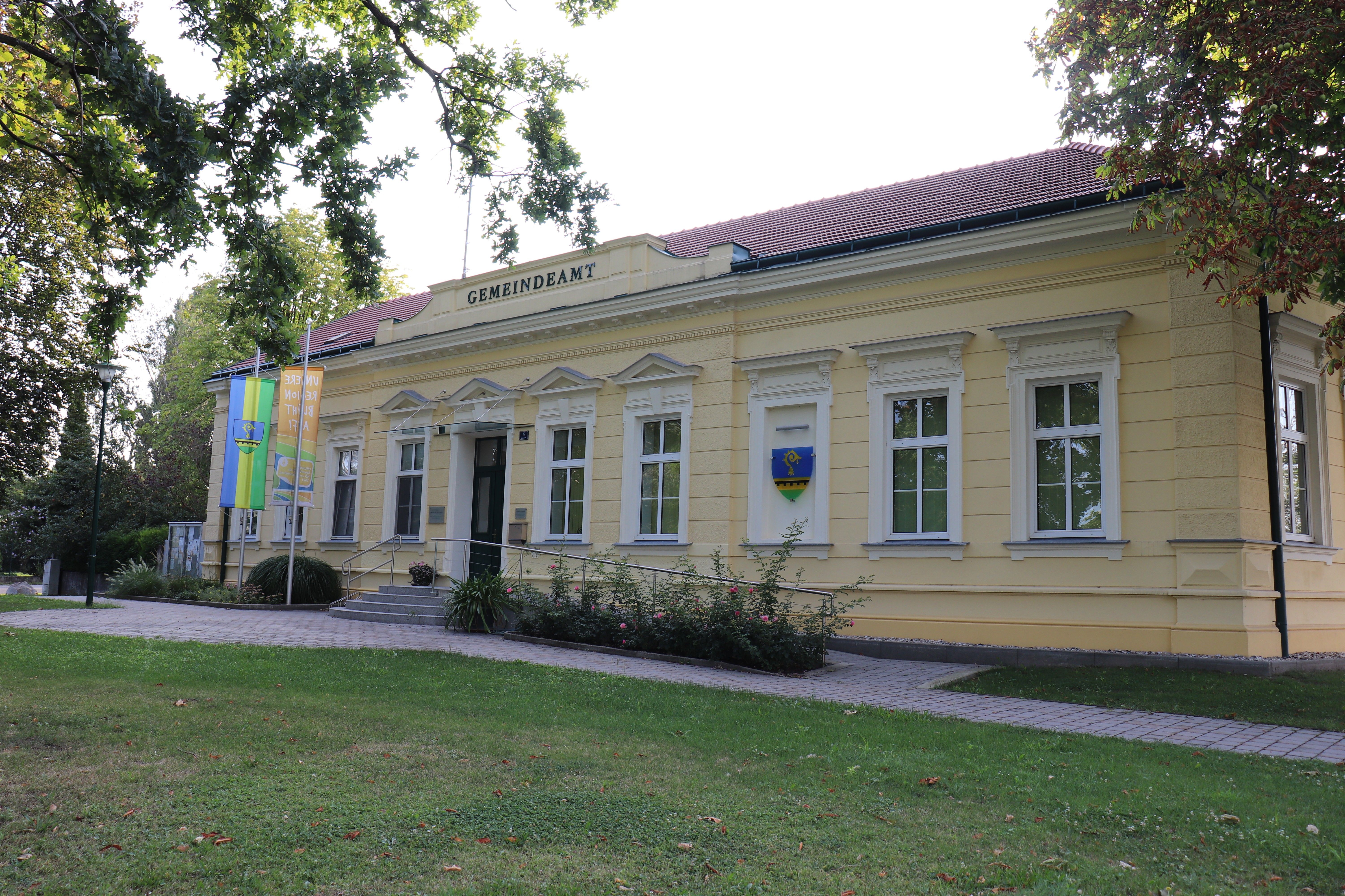 Raasdorf Town Hall, a bright administrative building with a red roof, flags, and a coat of arms, surrounded by trees and lawns.