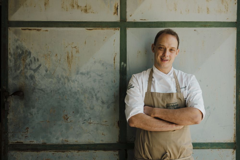 Cook in white uniform in front of rusty metal door, smiling into the camera.