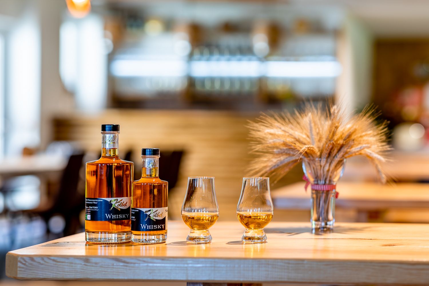 Two bottles of whisky and two glasses of whisky stand on a wooden table, with a bunch of cereals next to them.