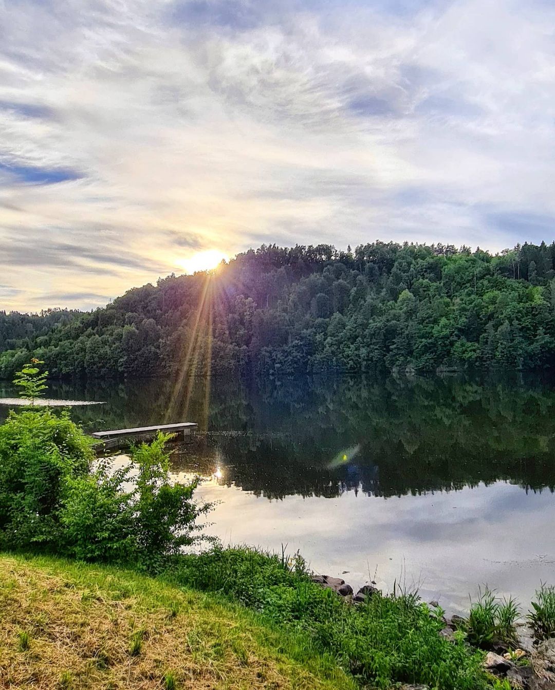 Blick auf das Wasser den Stausee Thurnberg mit einladendem Steg und dahinterliegendem Wald, hinter dem die Sonne hervorlugt. 