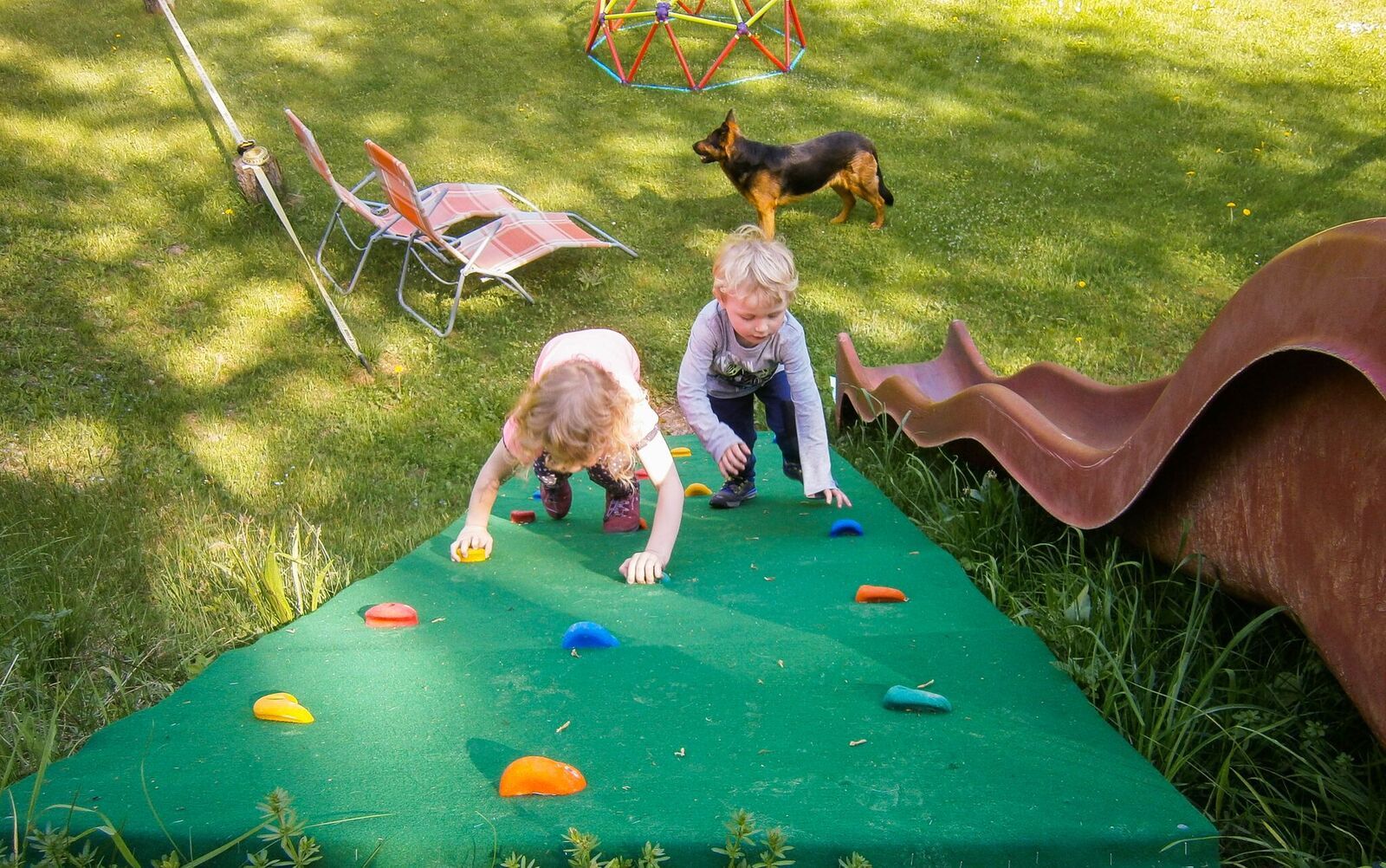 Two children climb up a green climbing wall on a playground, while a dog stands in the background.