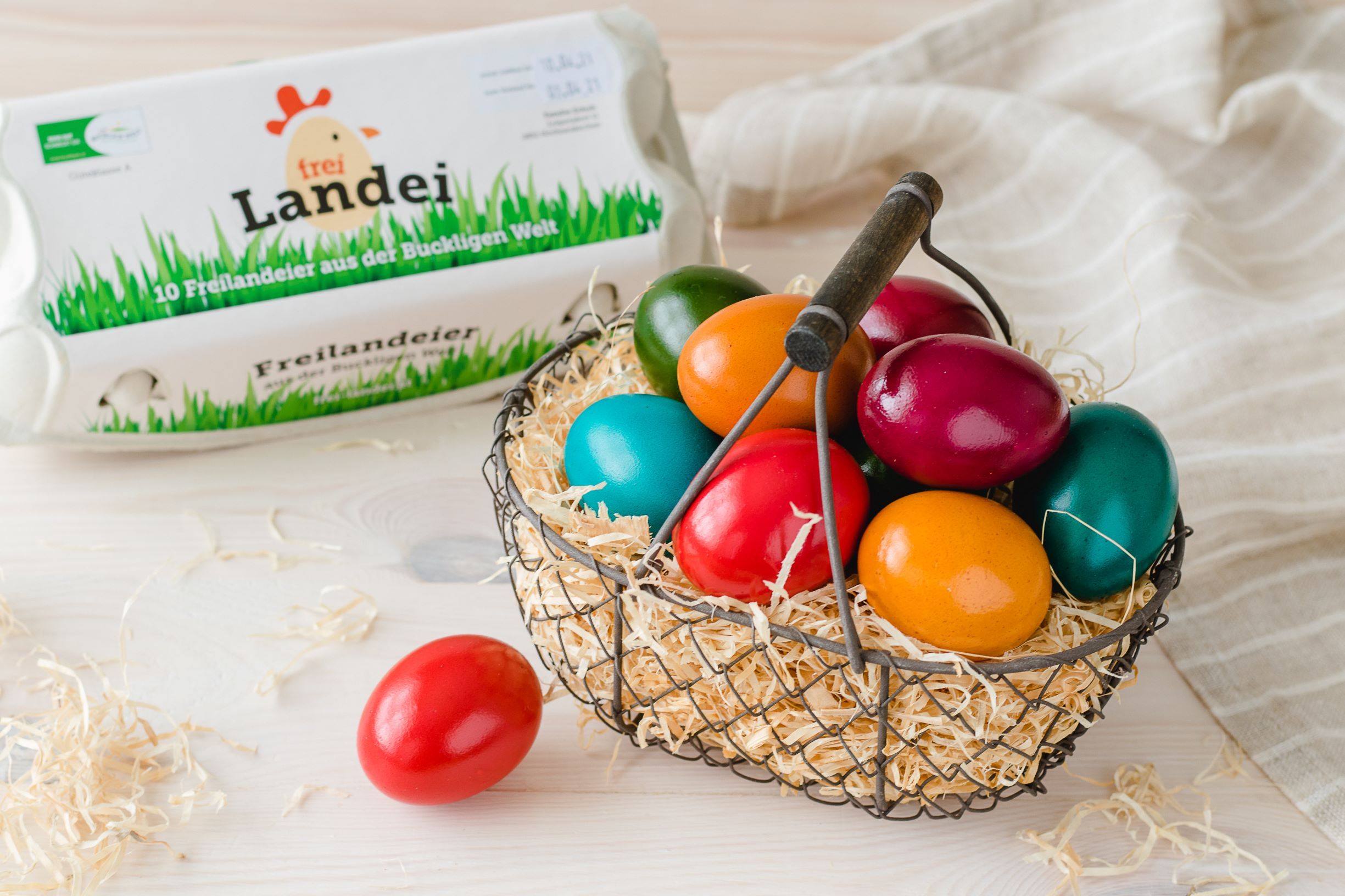 A basket filled with straw and colorful Easter eggs on a wooden table. Next to it, an egg carton, a red egg and a gray cloth.