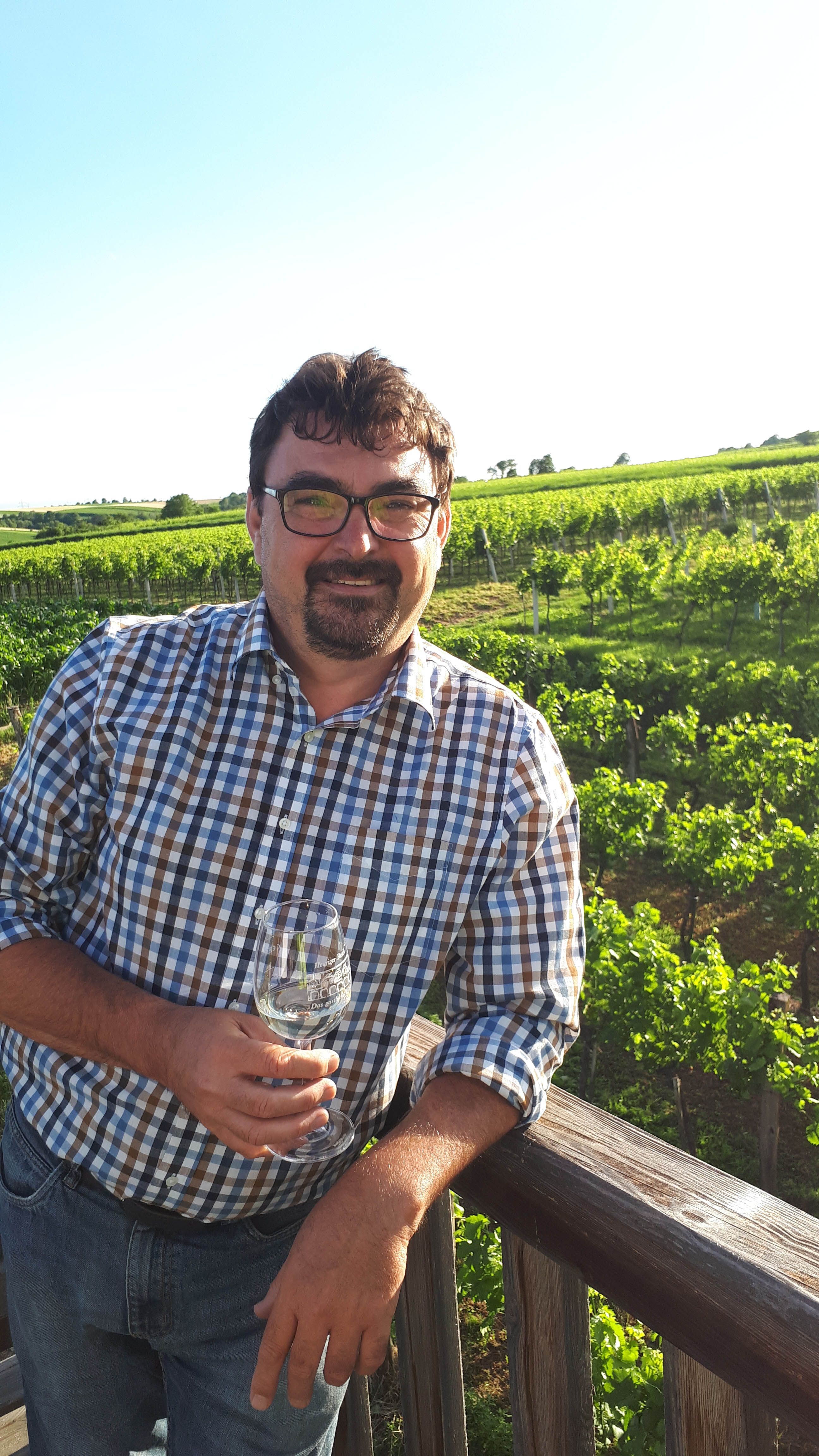 A man in a checked shirt stands in front of a vineyard with a wine glass in his hand.