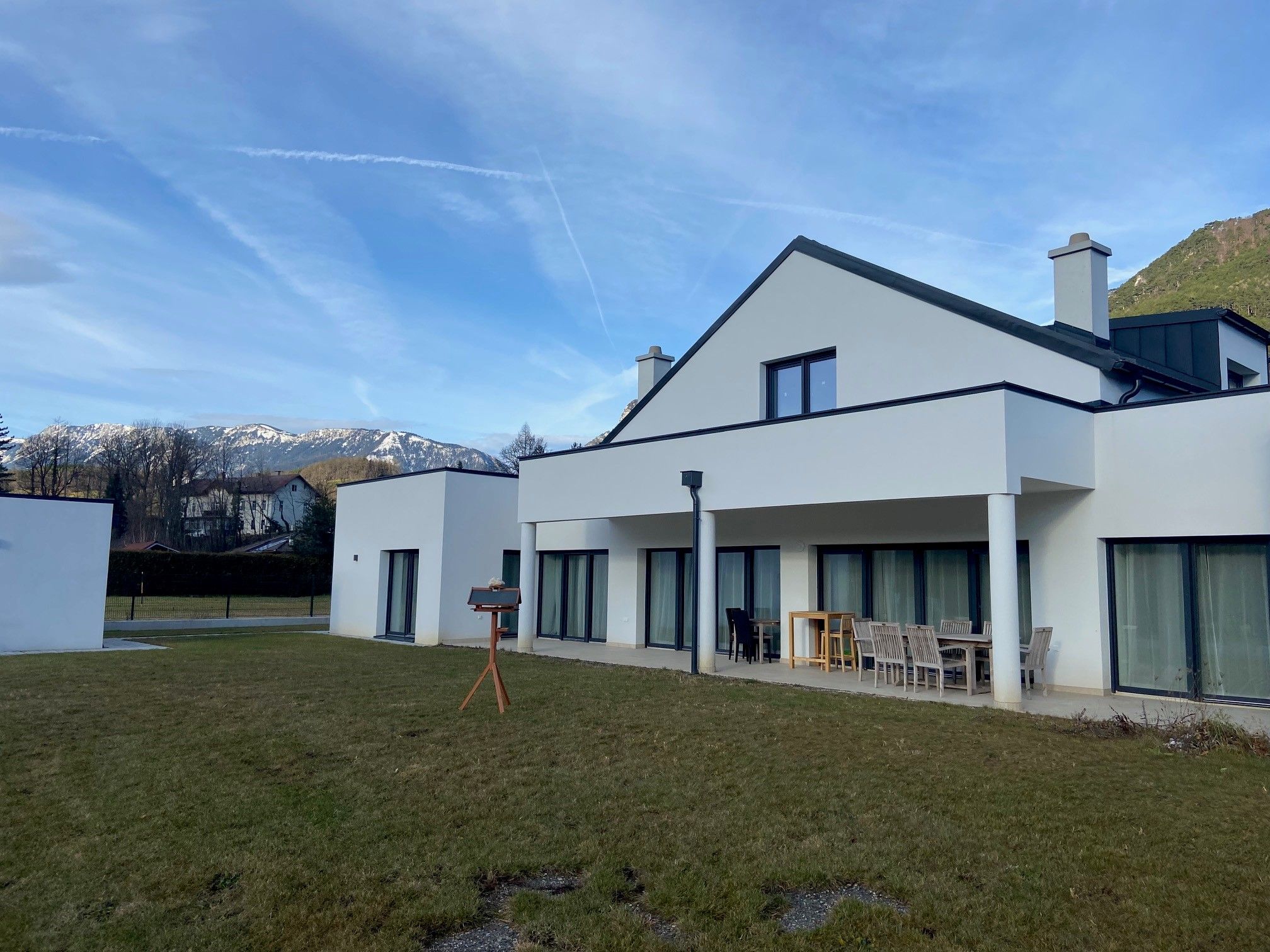 Modern white house with terrace and garden, snow-capped mountains in the background.