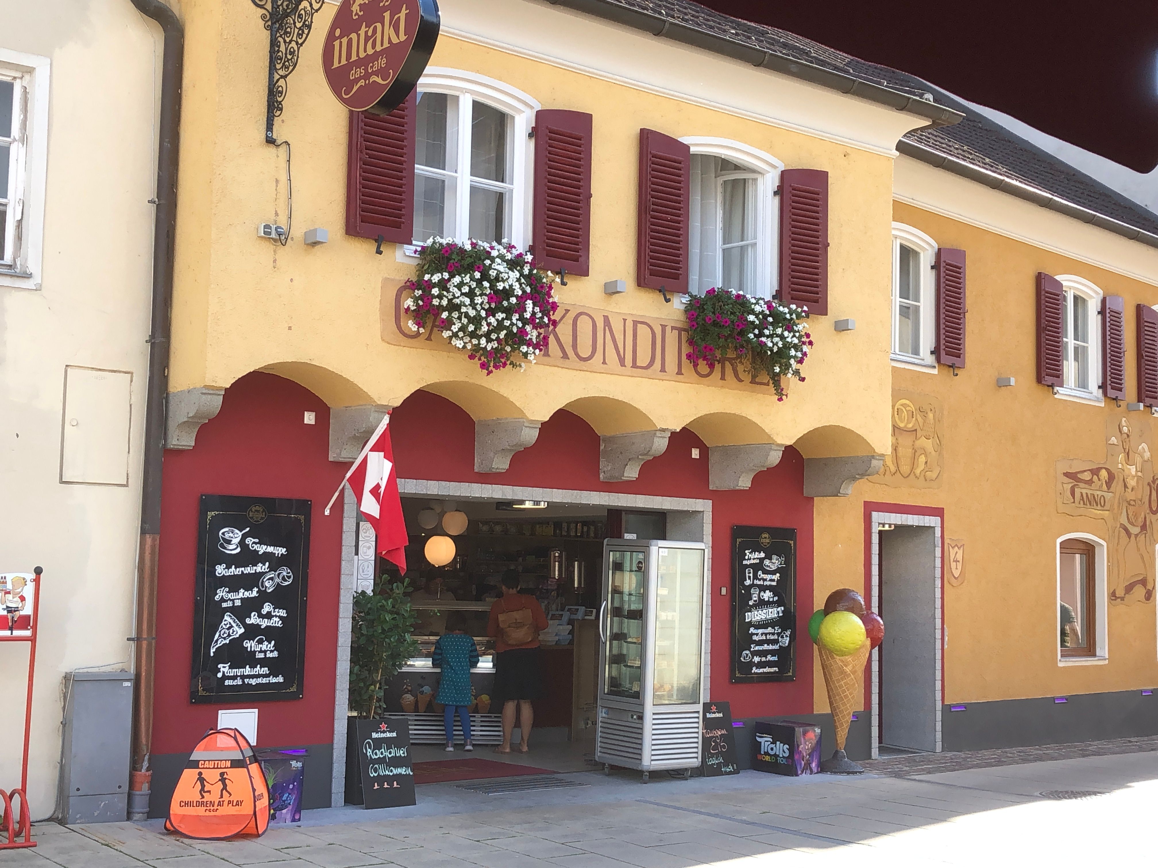 Exterior view of a café with a yellow façade, red shutters and floral decorations.