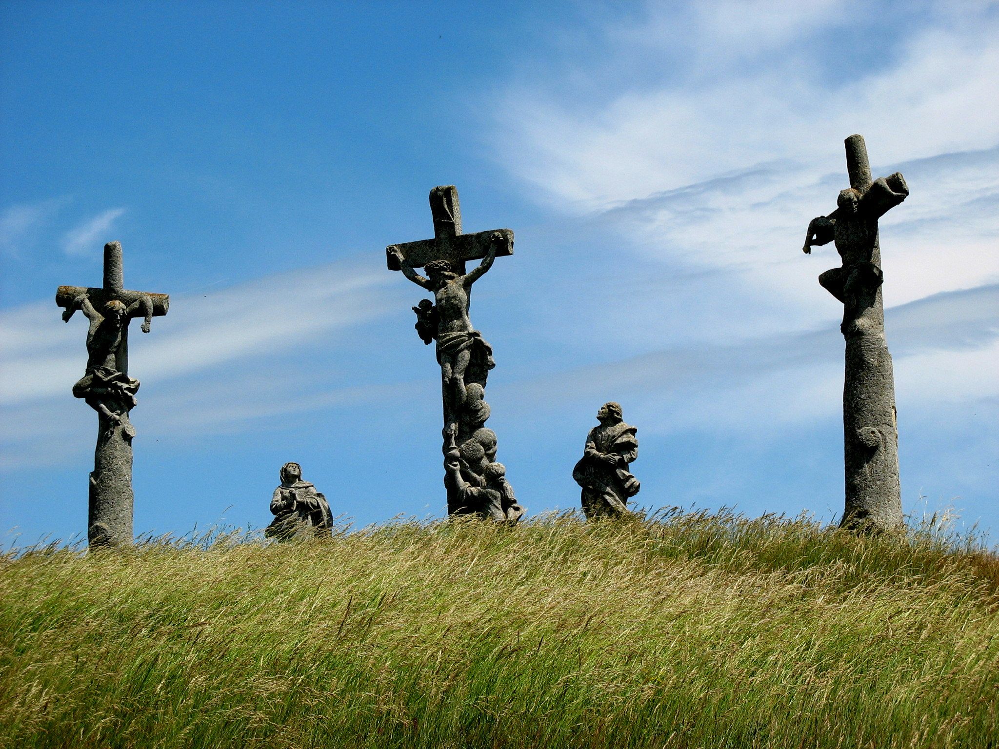Three crosses with figures on a hill under a blue sky.