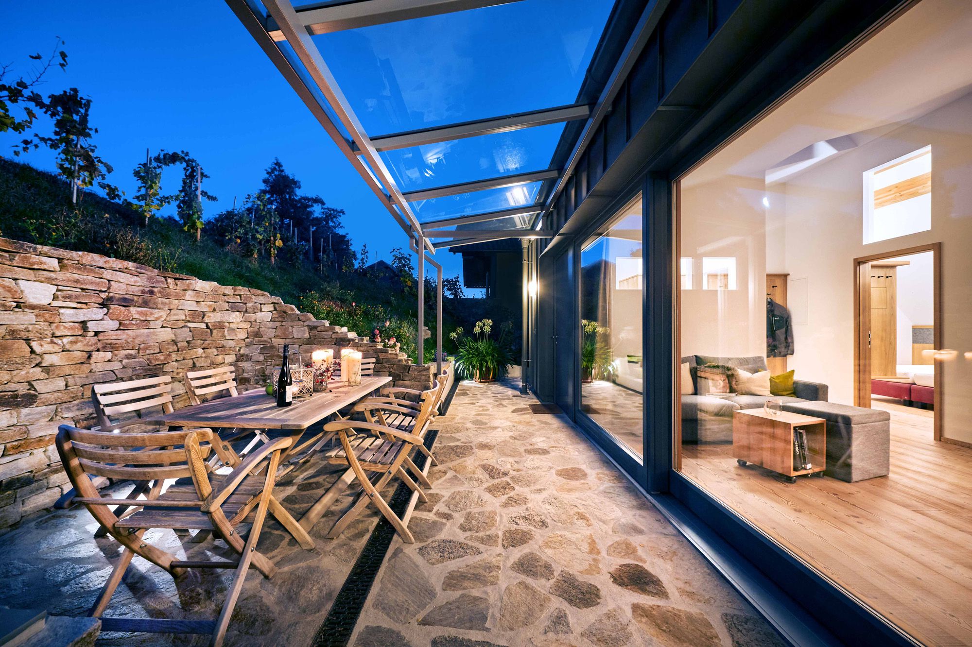 Terrace with wooden furniture and glass roof, view into the illuminated living room.