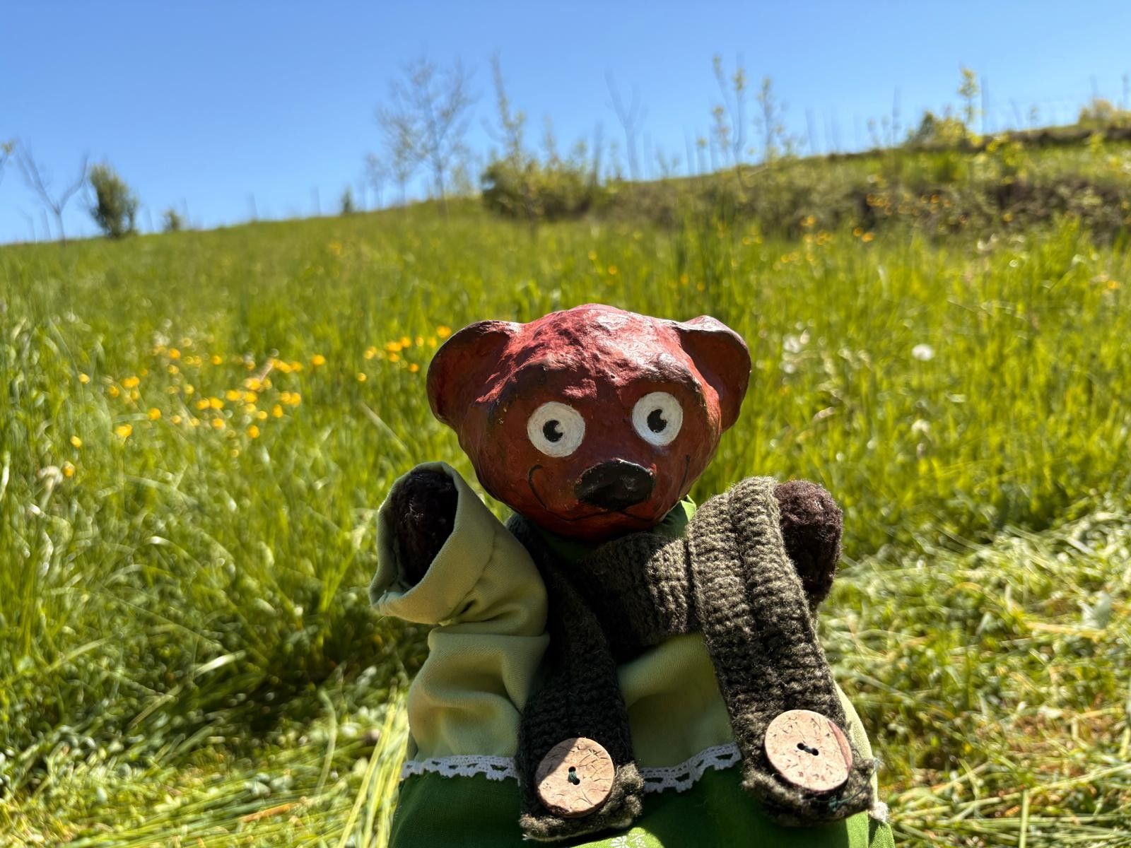 A handcrafted bear figurine with a red head and big eyes stands in a green meadow under a blue sky.
