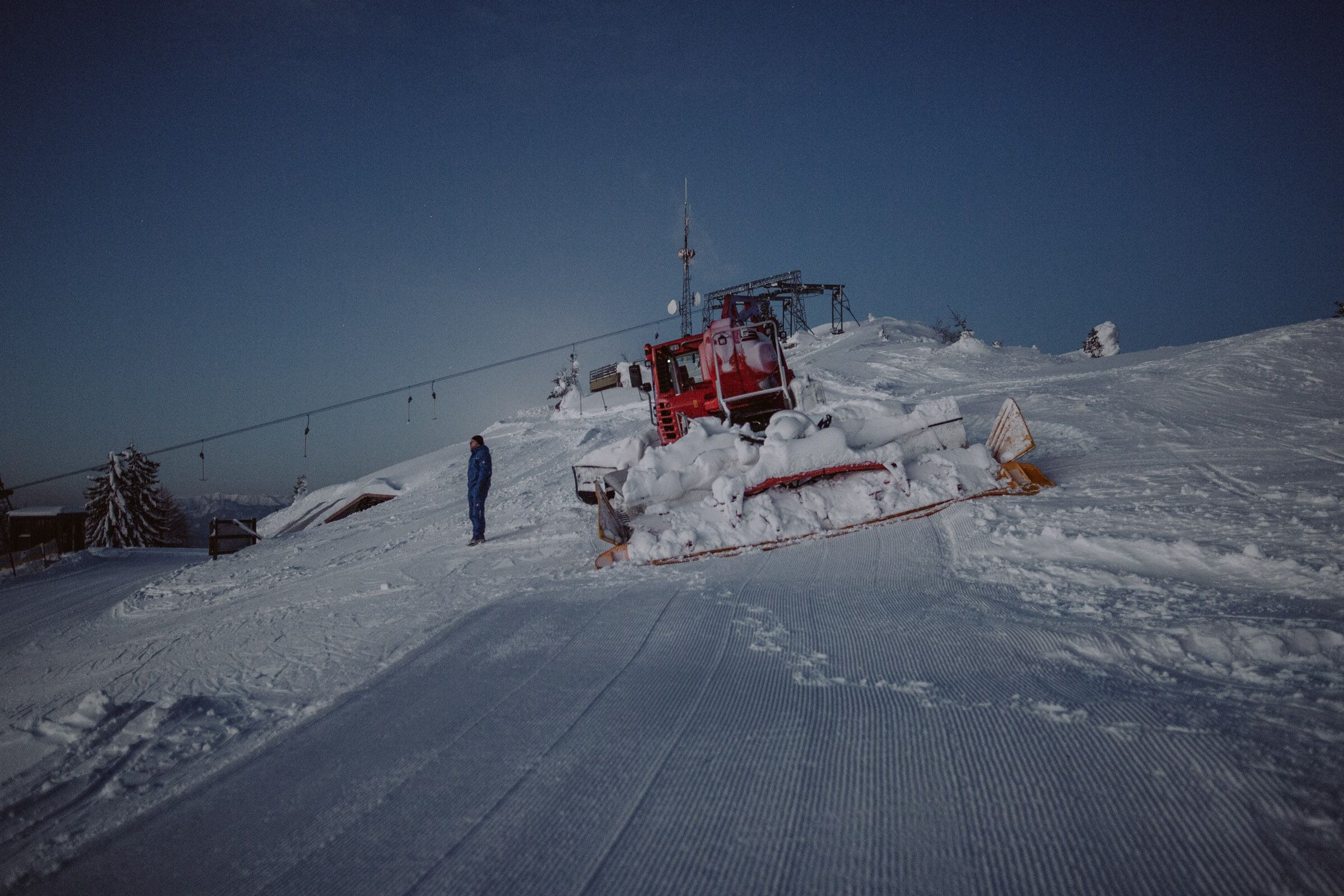 A snow groomer on a snow-covered ski slope at dusk in the Unterberg ski area.