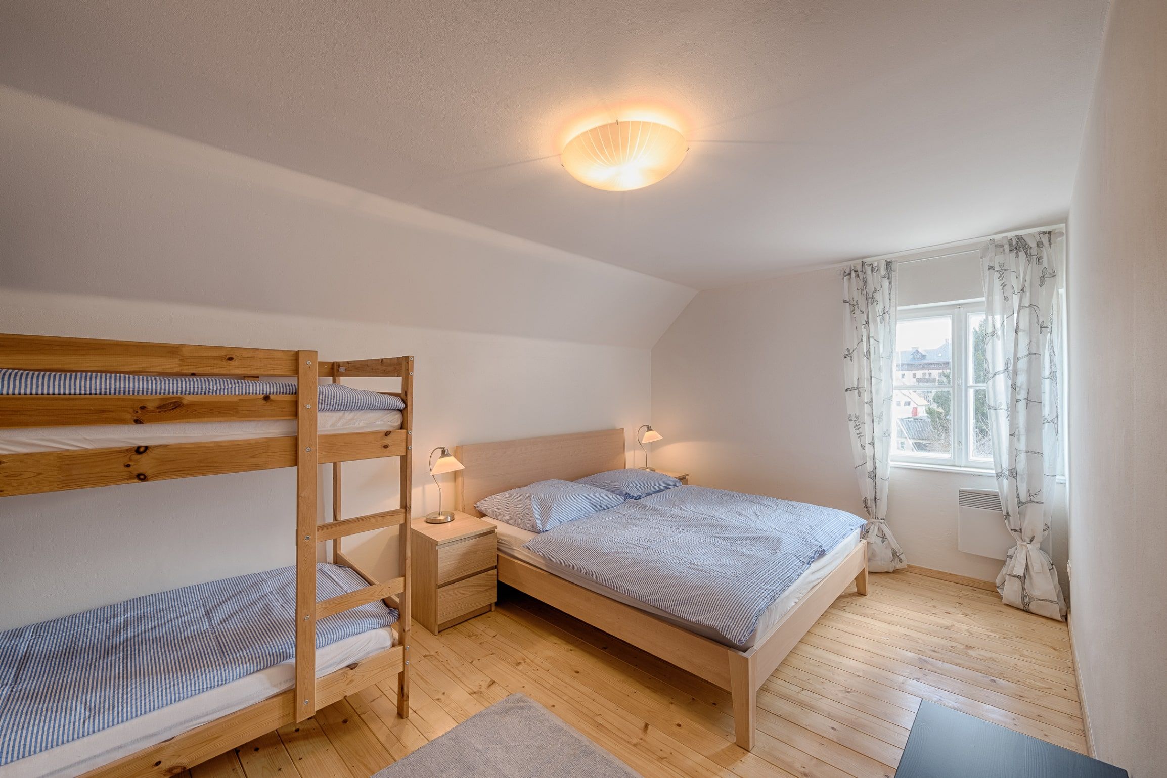 Bright bedroom with double bed and bunk bed, wooden floor and window with curtains.