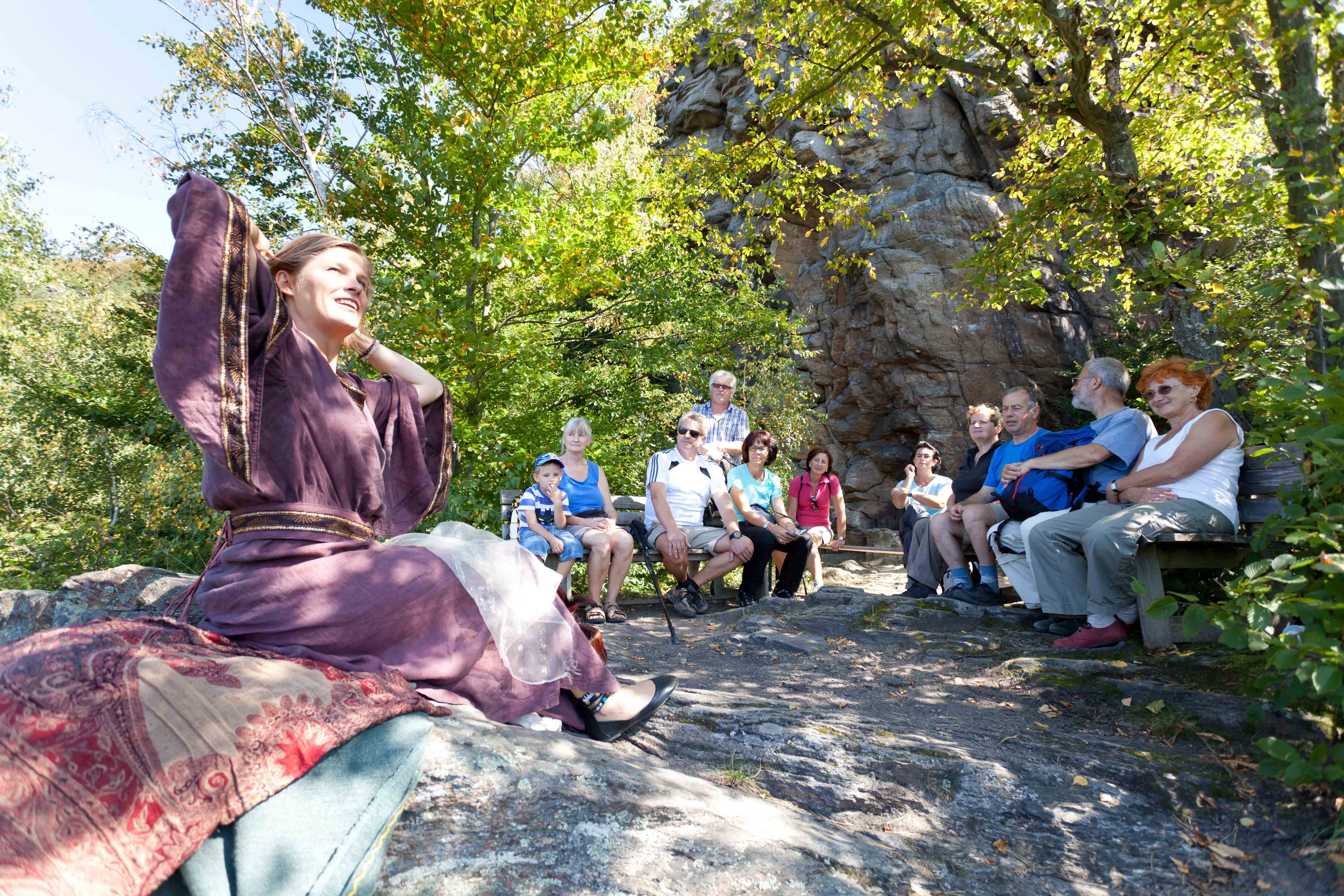 A woman in a purple robe tells a story to a group of people outside.