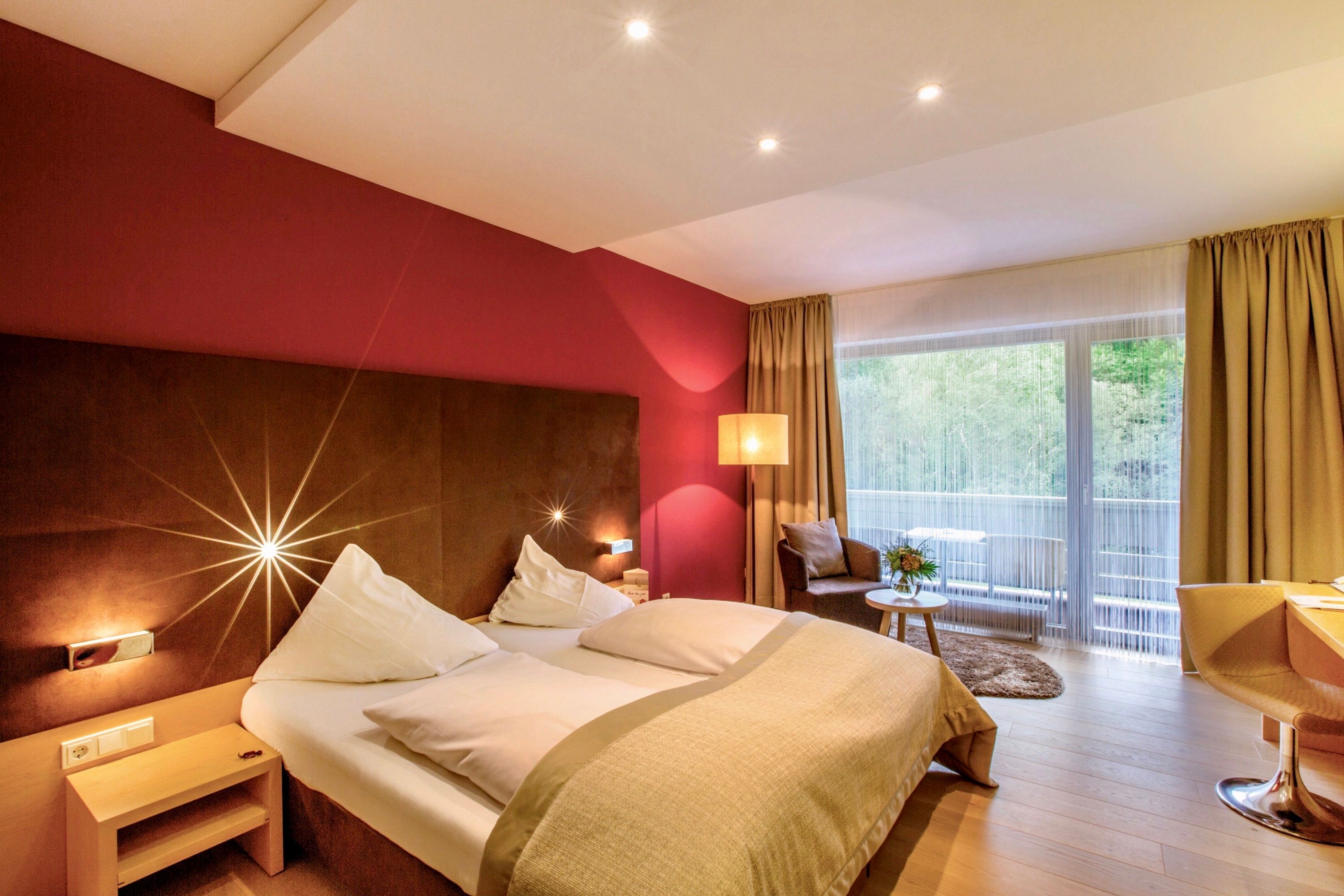 Modern hotel room with double bed, red wall, large window and seating area.