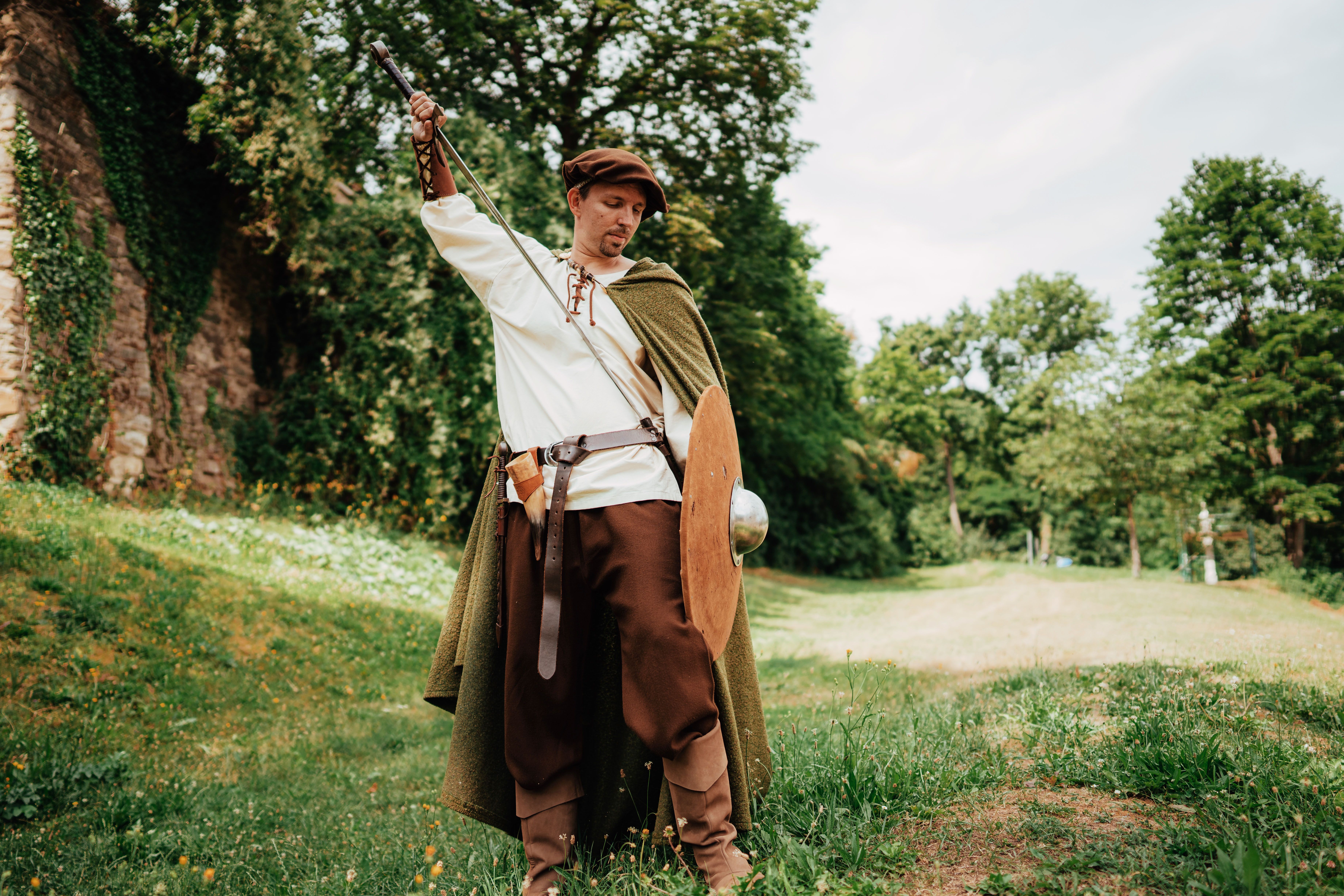 A man in medieval clothing with a sword and shield stands in a meadow in front of an overgrown stone wall.