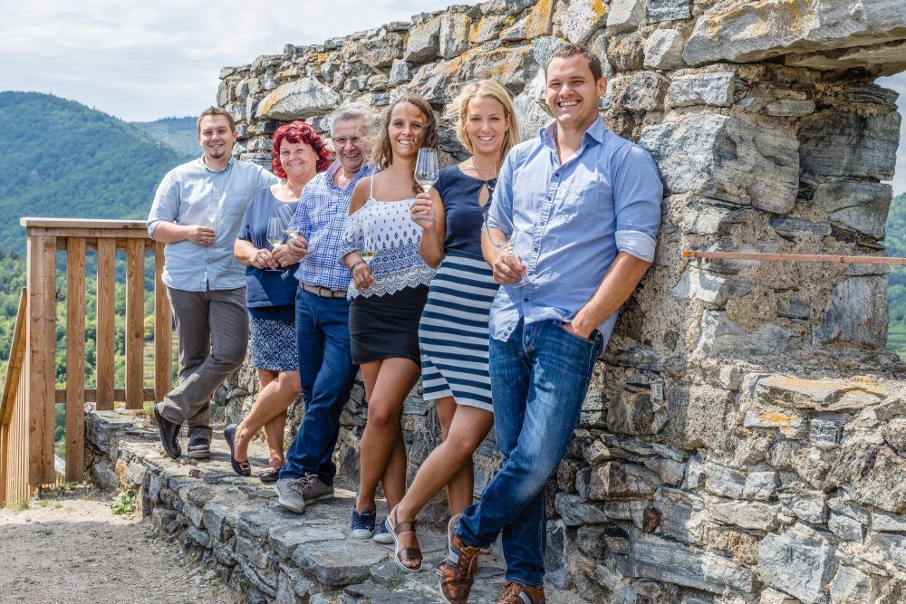 A group of six people stand smiling against a stone wall with glasses of wine in their hands, in front of a mountainous landscape.