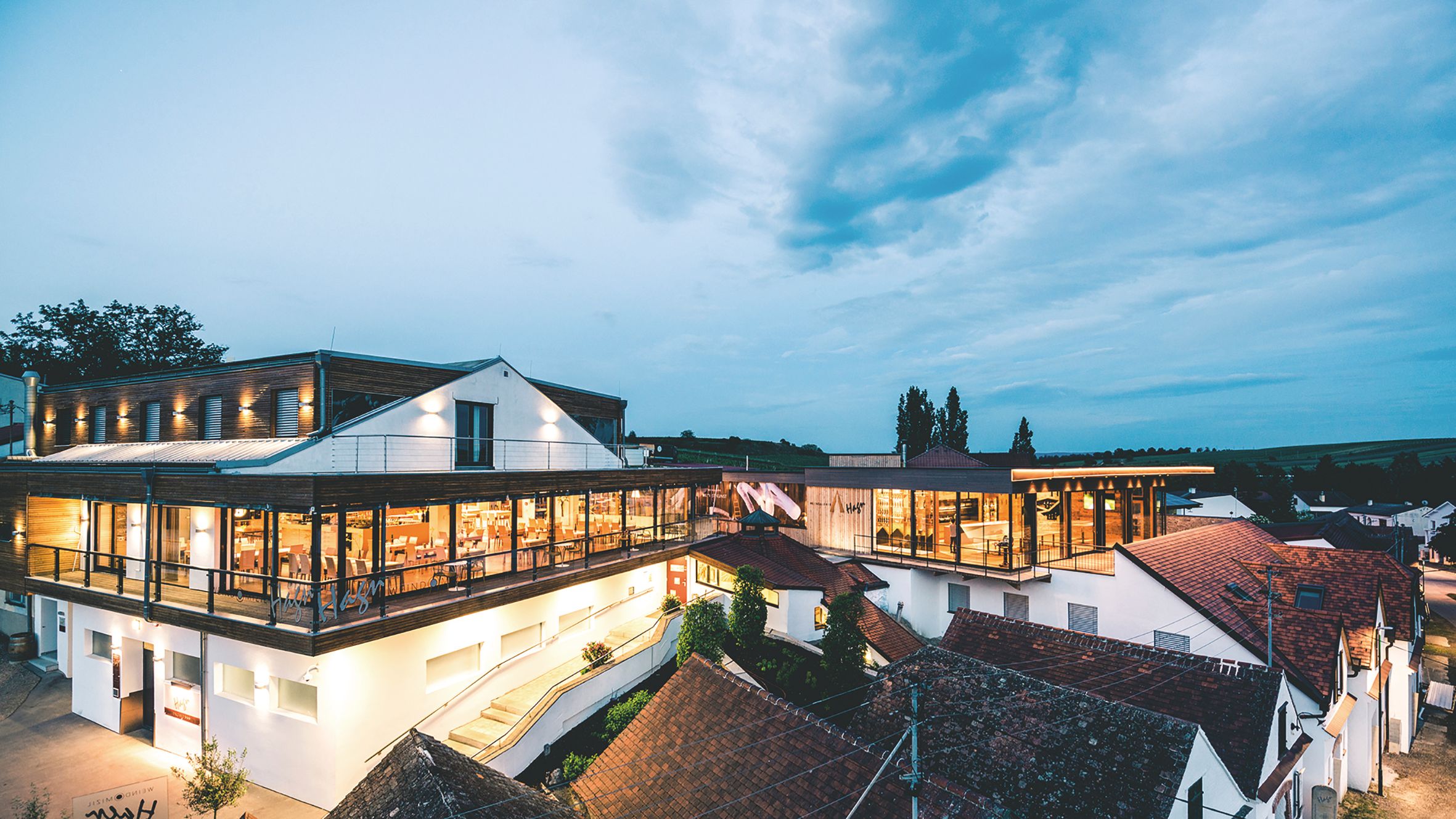 Modern winery architecture at dusk with illuminated interiors and surrounding buildings.