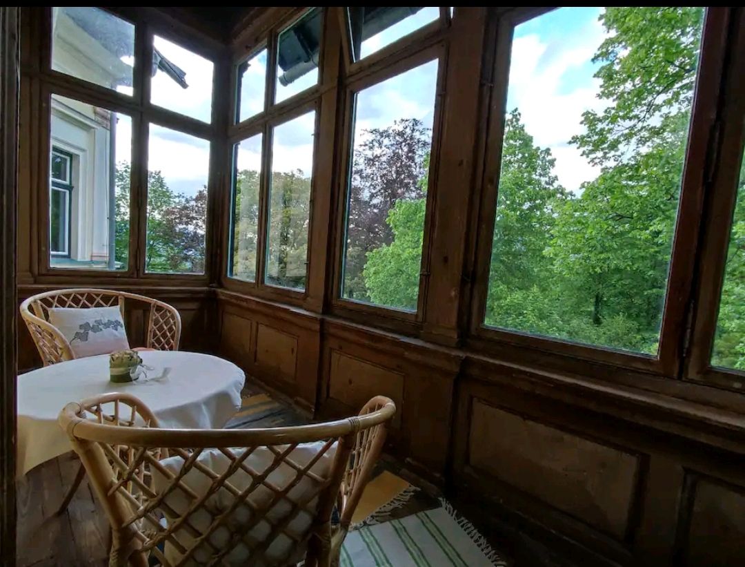 Veranda with table and two chairs, surrounded by large windows overlooking green trees.