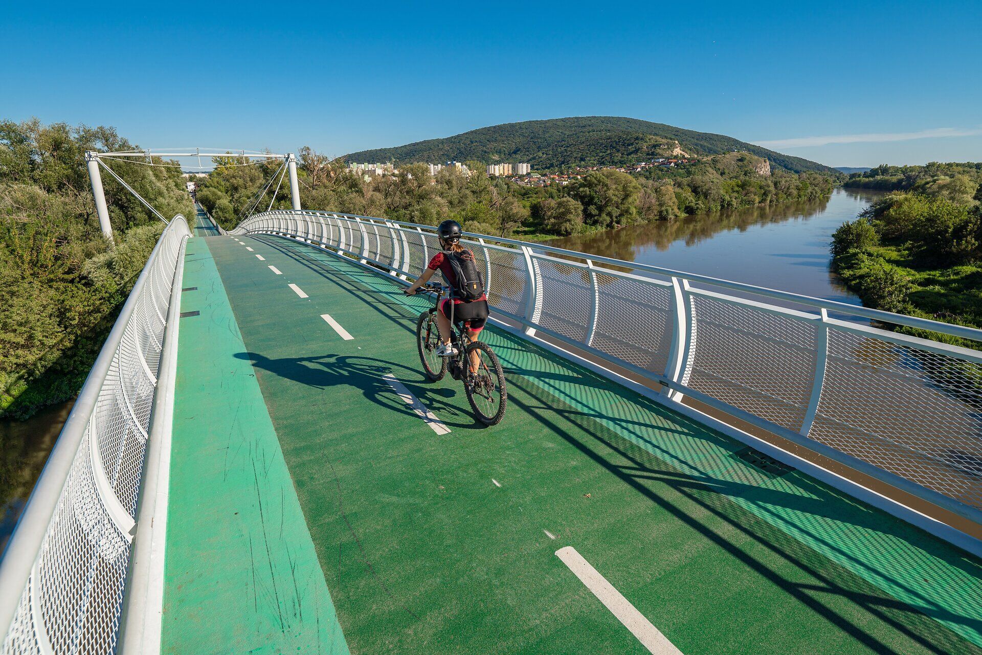 The Bridge of Freedom stretches majestically across the tranquil river, surrounded by lush greenery and rolling hills. Cyclists enjoy the fresh air and breathtaking views while experiencing the connection between nature and culture. A perfect place for an unforgettable view and relaxing hours in nature.